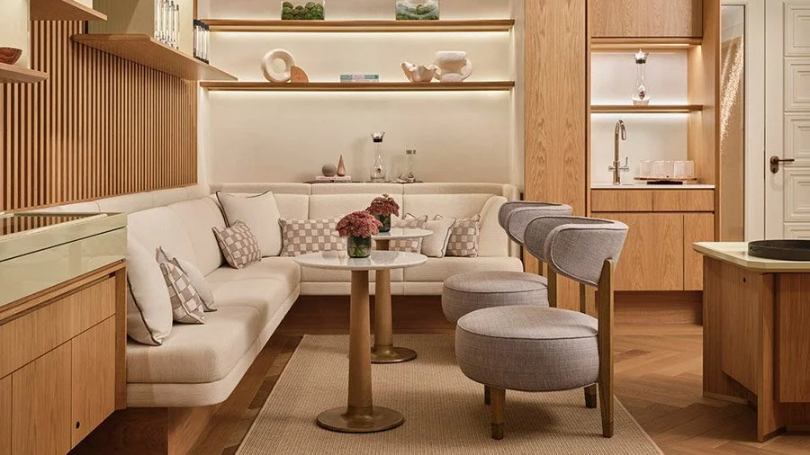 ein modernes Wohnzimmer mit einer weißen Couch, mehreren Kissen, einem runden Couchtisch mit Blumen, grauen Stühlen, Holzregalen mit Deko-Objekten und einem kleinen Bereich mit Spüle im Hintergrund.