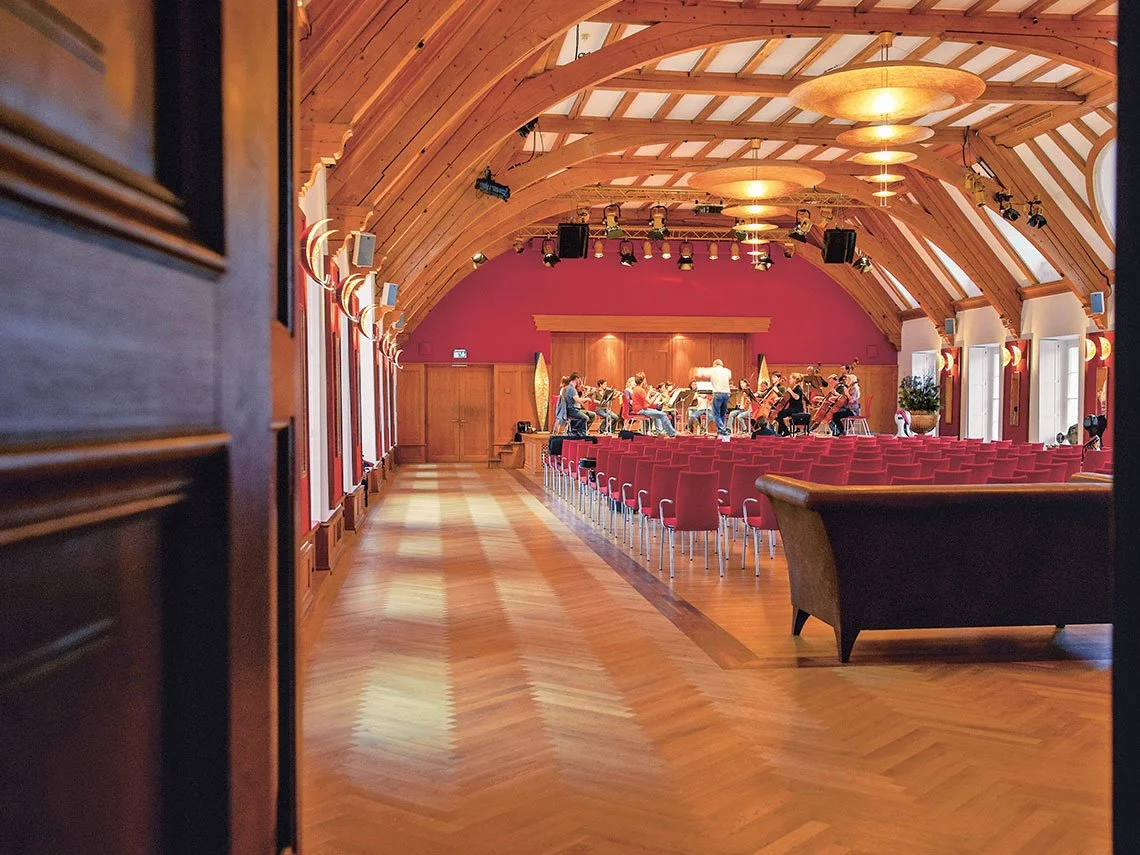 Ein Saal mit hohen Holzgewölbedecken, rotem Wänden und Reihen von Stühlen, auf einer Bühne spielen Musiker mit einem Dirigenten.