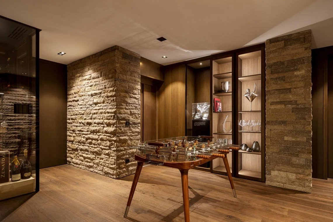 Innenraum mit einem Flipper-Automaten, Steinsäulen und Einbauschränken aus Holz, dekorativen Objekten und Weinflaschen.