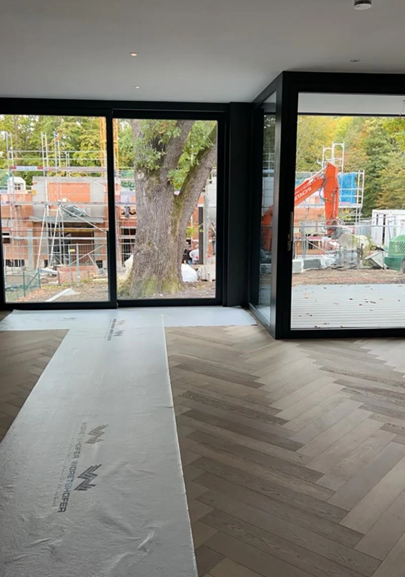 Innenansicht eines Hauses unter Bau, mit großen Fensterfronten, Blick auf eine Baustelle mit Gerüsten, Bauarbeiten und einem Baumaschinen im Außenbereich