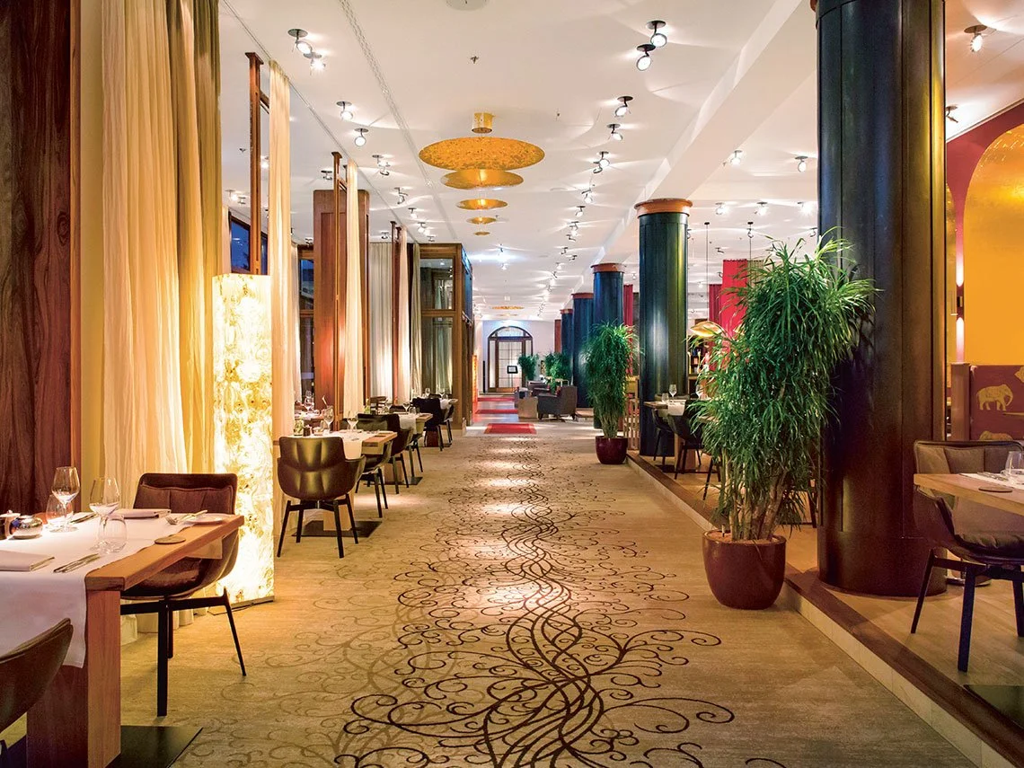 Innenansicht eines eleganten Restaurants mit Tischen, Stühlen, Pflanzen, dunklen Säulen und warmem Licht.