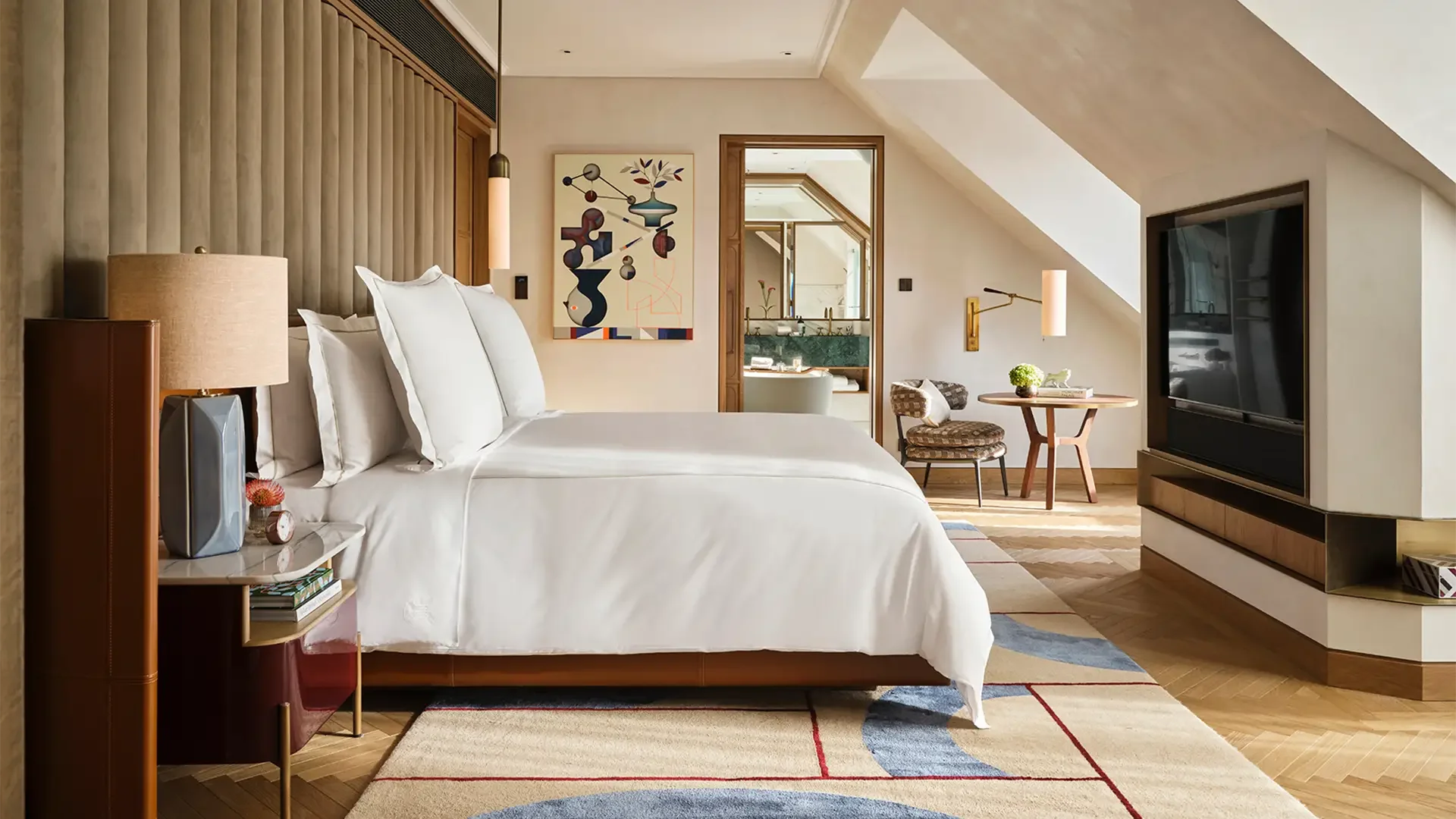 Ein Hotelzimmer mit einem großen Bett, Nachttisch mit Lampe, moderner Kunst an der Wand, Fernseher, Sitzbereich mit Stuhl und Tisch, und Badezimmer im Hintergrund.