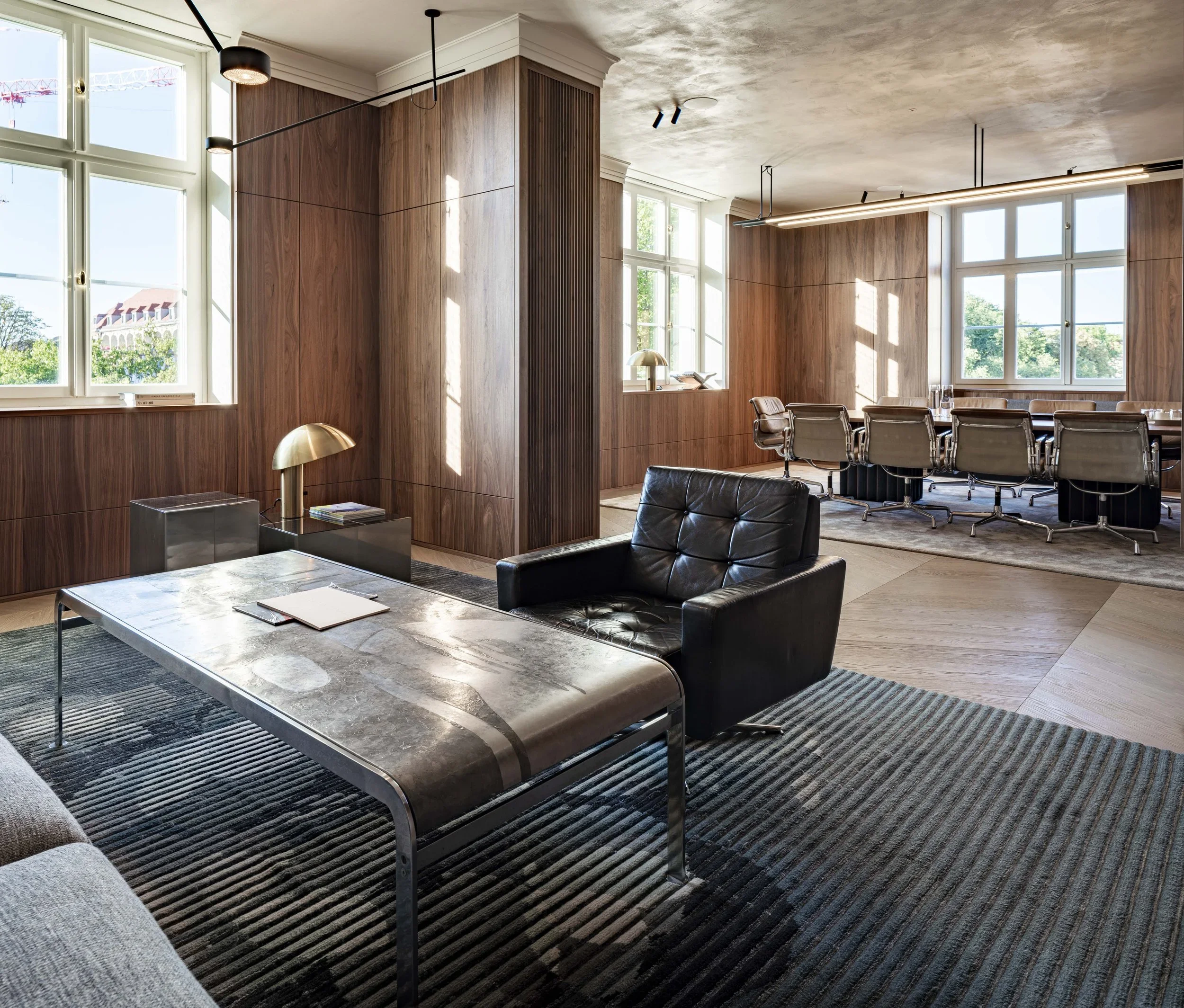 Innenansicht eines modernen Büros mit Holzvertäfelungen, großen Fenstern, einem schwarzen Ledersessel, einem Konferenztisch mit Stühlen und einem Schreibtisch.
