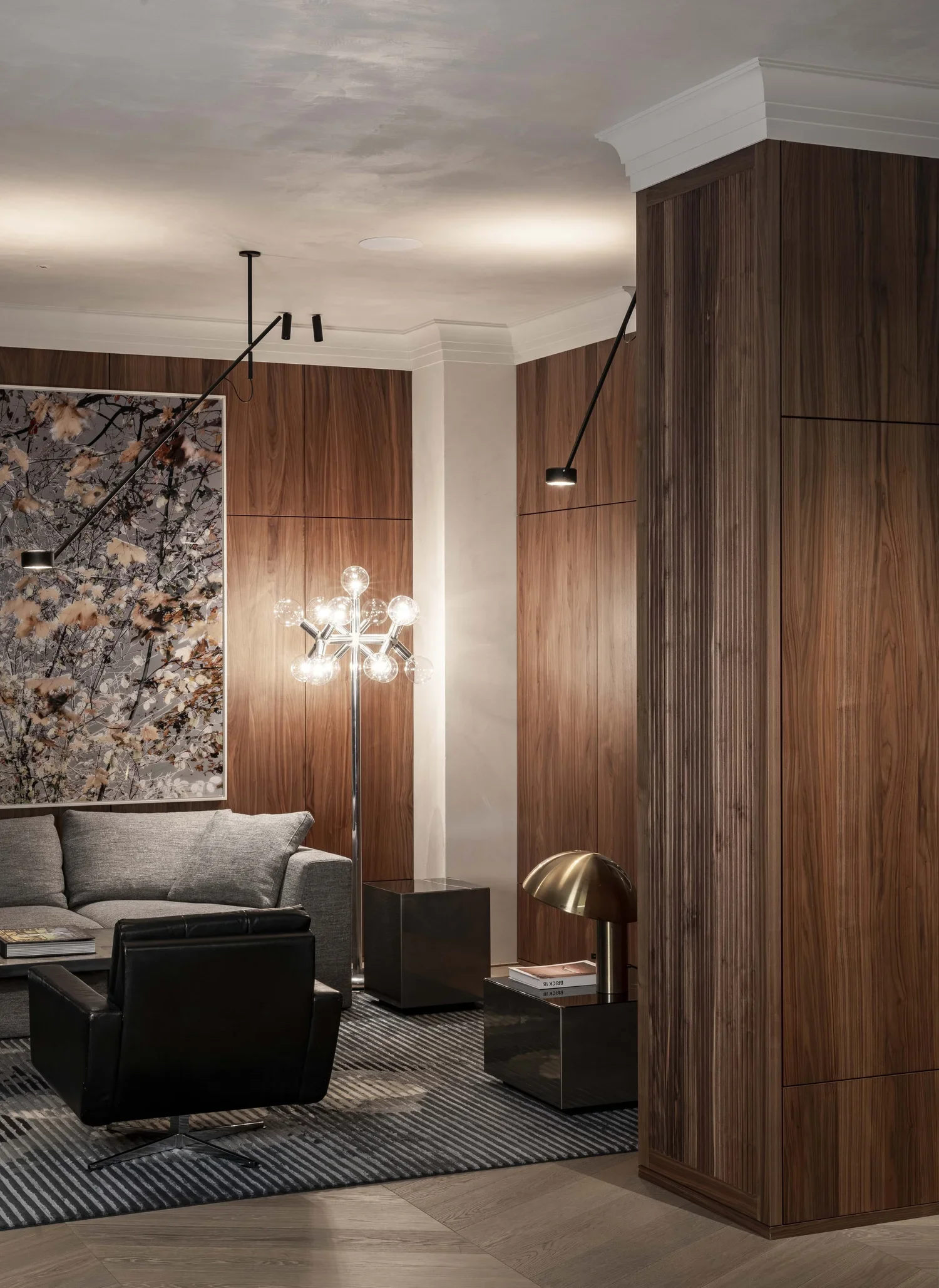 Moderner Wohnzimmerbereich mit grauer Couch, schwarzer Sessel, Holz-Wandverkleidung, Stehlampe mit mehreren Glühbirnen und Deckenleuchten.