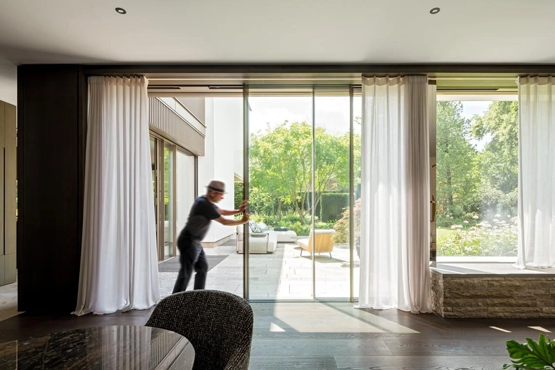 Ein Mann öffnet eine große Glastür, die in einen Garten mit Bäumen und Sitzmöbeln führt, während die Innenseite des Hauses mit dunklem Holz, weißen Vorhängen und einem Esstisch sichtbar ist.