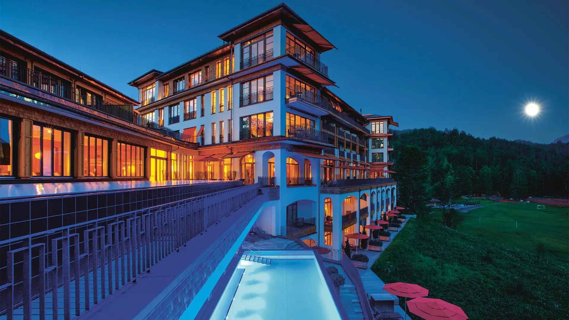 Ein modernes Hotelgebäude bei Nacht mit beleuchteten Fenstern, einem beheizten Pool im Vordergrund, grünem Rasen und Bäumen im Hintergrund sowie einem hellen Vollmond am Himmel.