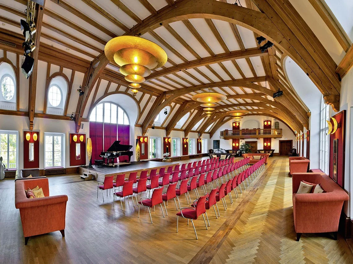 Ein Konzertsaal mit einem Flügel auf der Bühne, Sitzreihen in Rot, gemütliche Sitzbänke an den Wänden, große Fenster, Holzdecke mit Armaturen, dekorative Wandlampen.