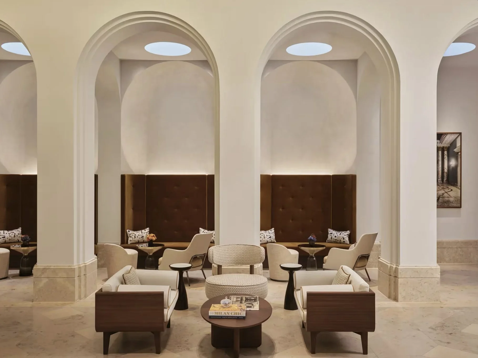 Eleganter Hotel-Lounge mit weißen Sesseln, dunkelbraunen Lederkopfhockern, kleinen Tischen, einem runden Ohrensessel und einer Stehlampe, in einem Raum mit hohen Decken und runden Lichtöffnungen.