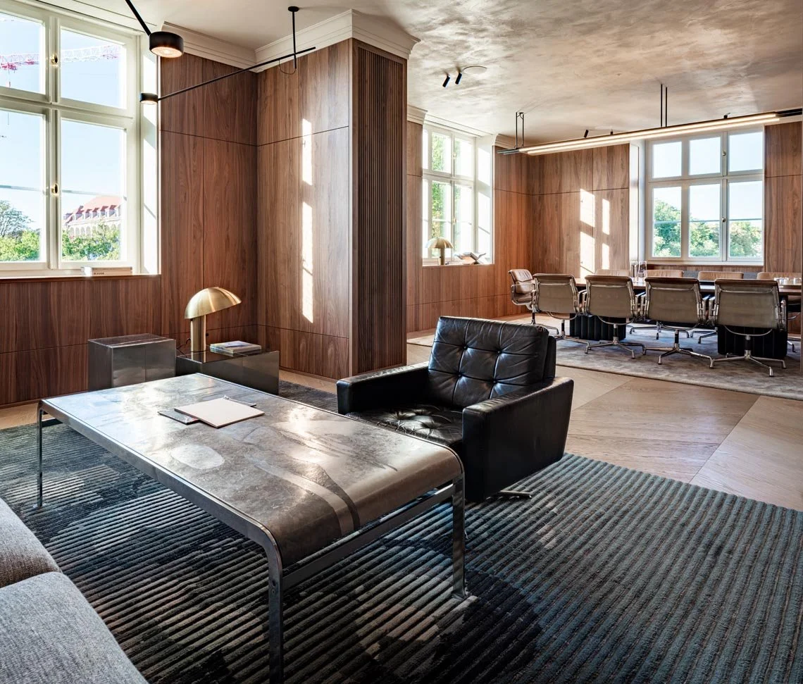 Ein modernes, lichtdurchflutetes Konferenzzimmer mit Holzdekoren, einem großen Tisch mit Stühlen im Hintergrund, einem schwarzen Ledersessel, einem Marmortisch, einem goldfarbenen Schreibtischlampe und großen Fenstern, die auf eine grüne Landschaft b