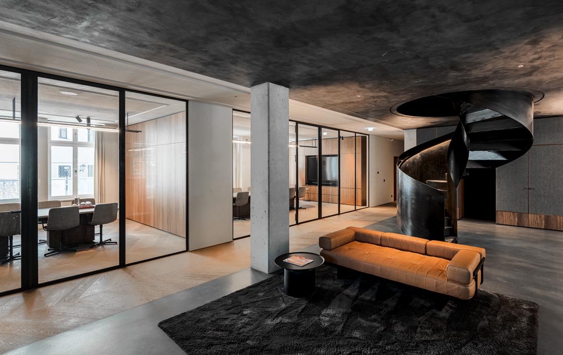 Modernes Wohnzimmer mit braunem Sofa, schwarzem Teppich, spiralförmiger schwarzen Treppe, Glastrennung und Blick auf einen hellen Arbeitsraum.