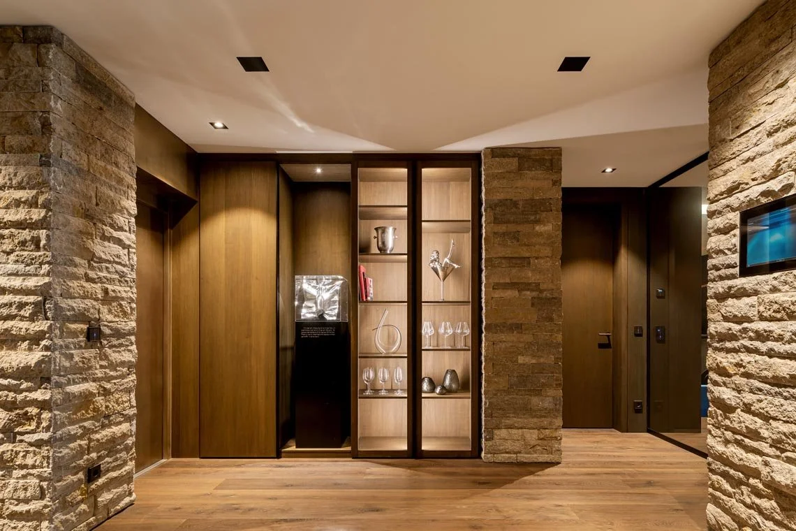 Innenansicht eines modernen Wohnzimmers mit Holzwänden, Steinsäulen und offenen Regalen mit Glaswaren und Dekorationen.