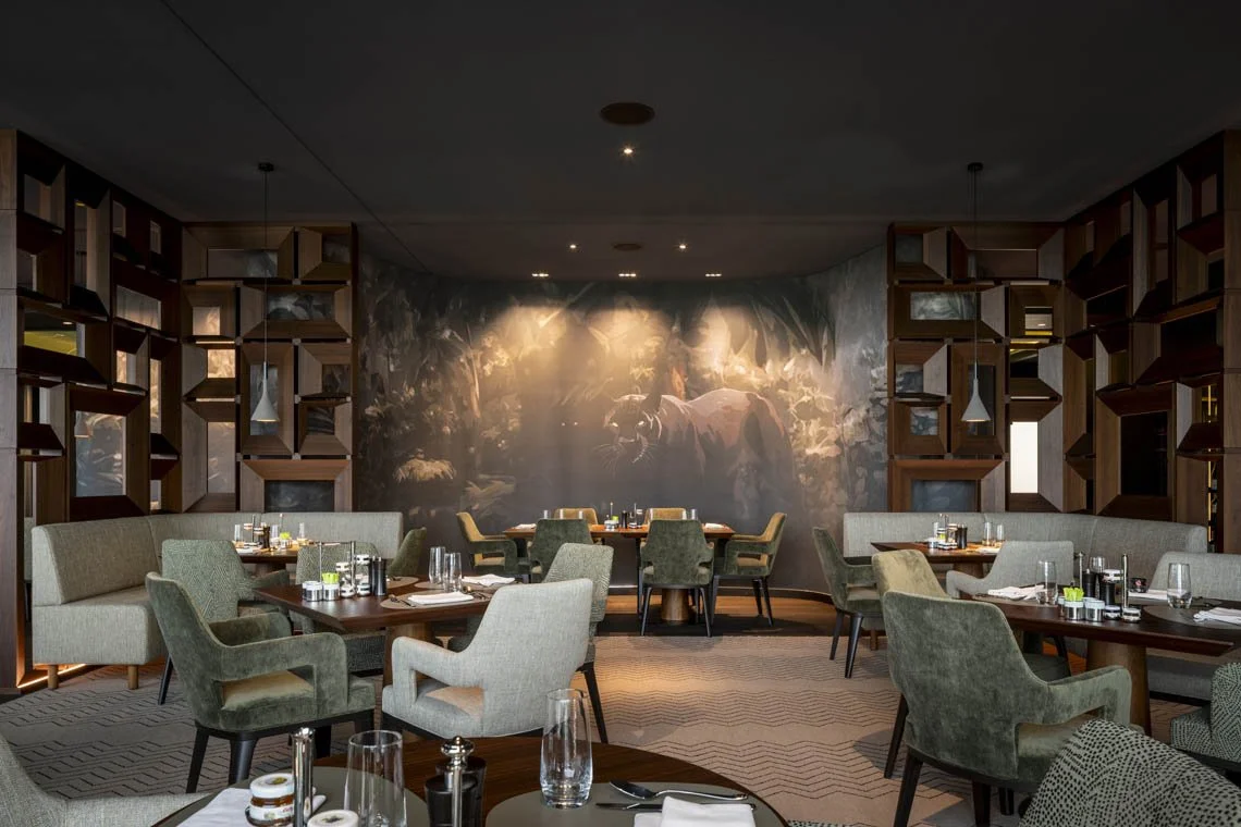 Ein stilvoll eingerichtetes Restaurant mit grünen und beigen Sesseln, Holztischen und einer Wand mit einem Wandbild eines Tigers in einem Dschungel.
