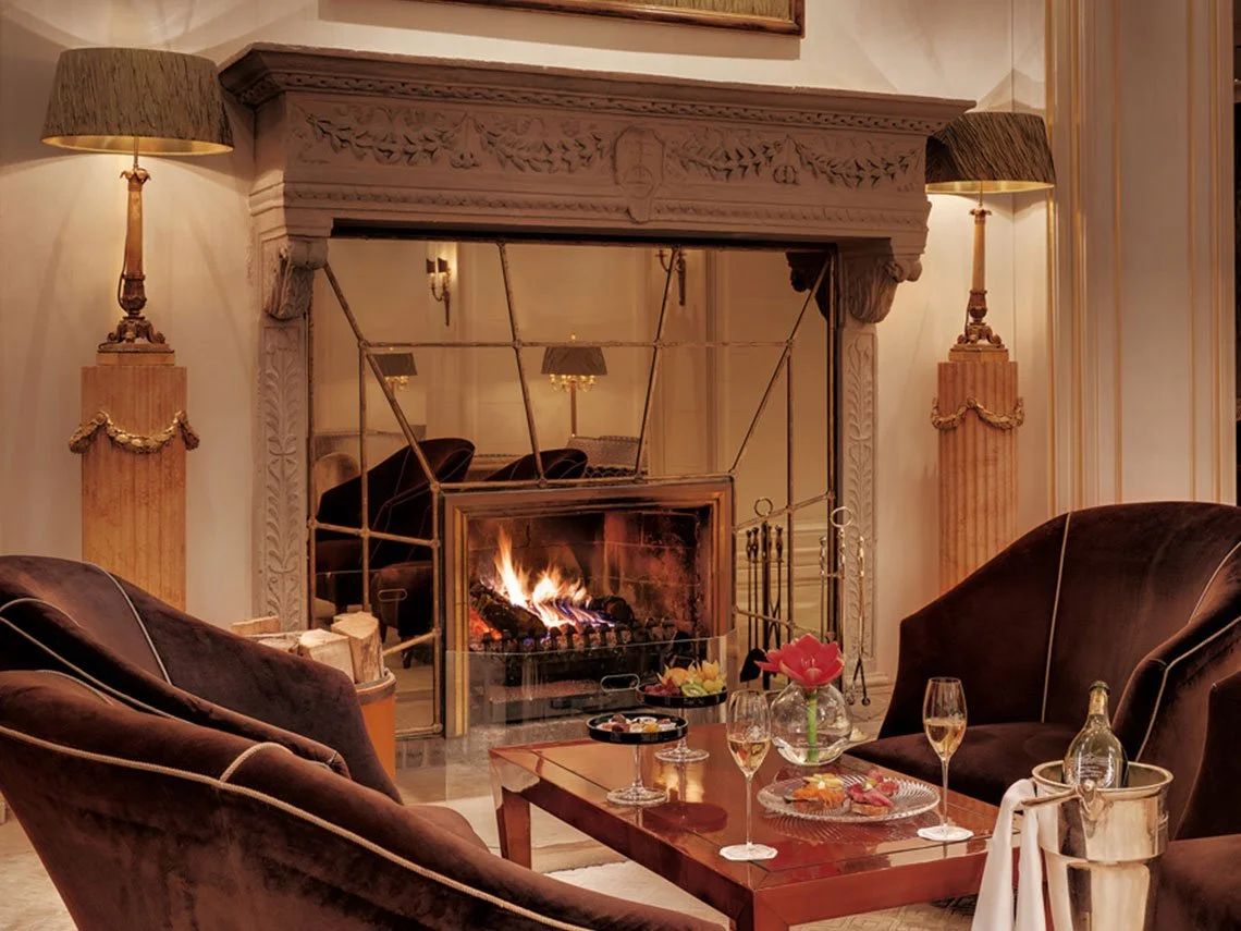 Ein gemütliches Wohnzimmer mit einem Kamin, zwei dunklen Sofas, einem Holztisch mit Getränken und Snacks, Blumen und Stehlampen.