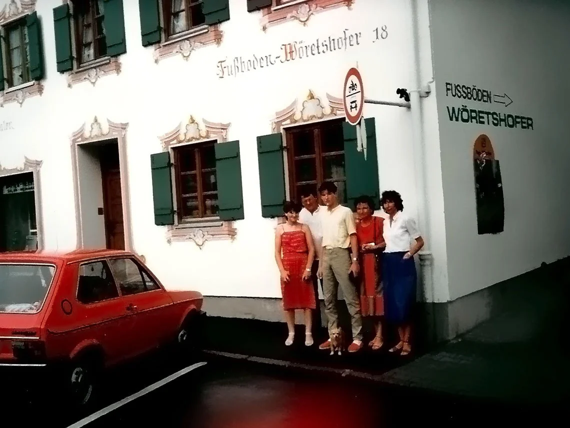 Fünf Menschen stehen vor einem weißen Gebäude mit grünen Fensterläden, die Schmiedekunst hergestellt haben, während ein rotes Auto im Vordergrund geparkt ist.