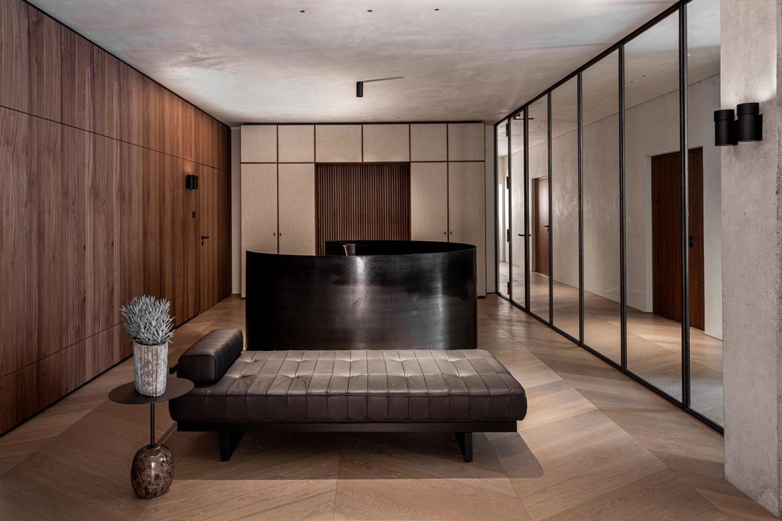 Modernes Innenraum mit Holz- und Betonwänden, einem schwarzen Empfangstresen, einem dunkelbraunen Lounge-Sessel, einem Beistelltisch mit Pflanze und Glasvase, sowie gläsernen Trennwänden.