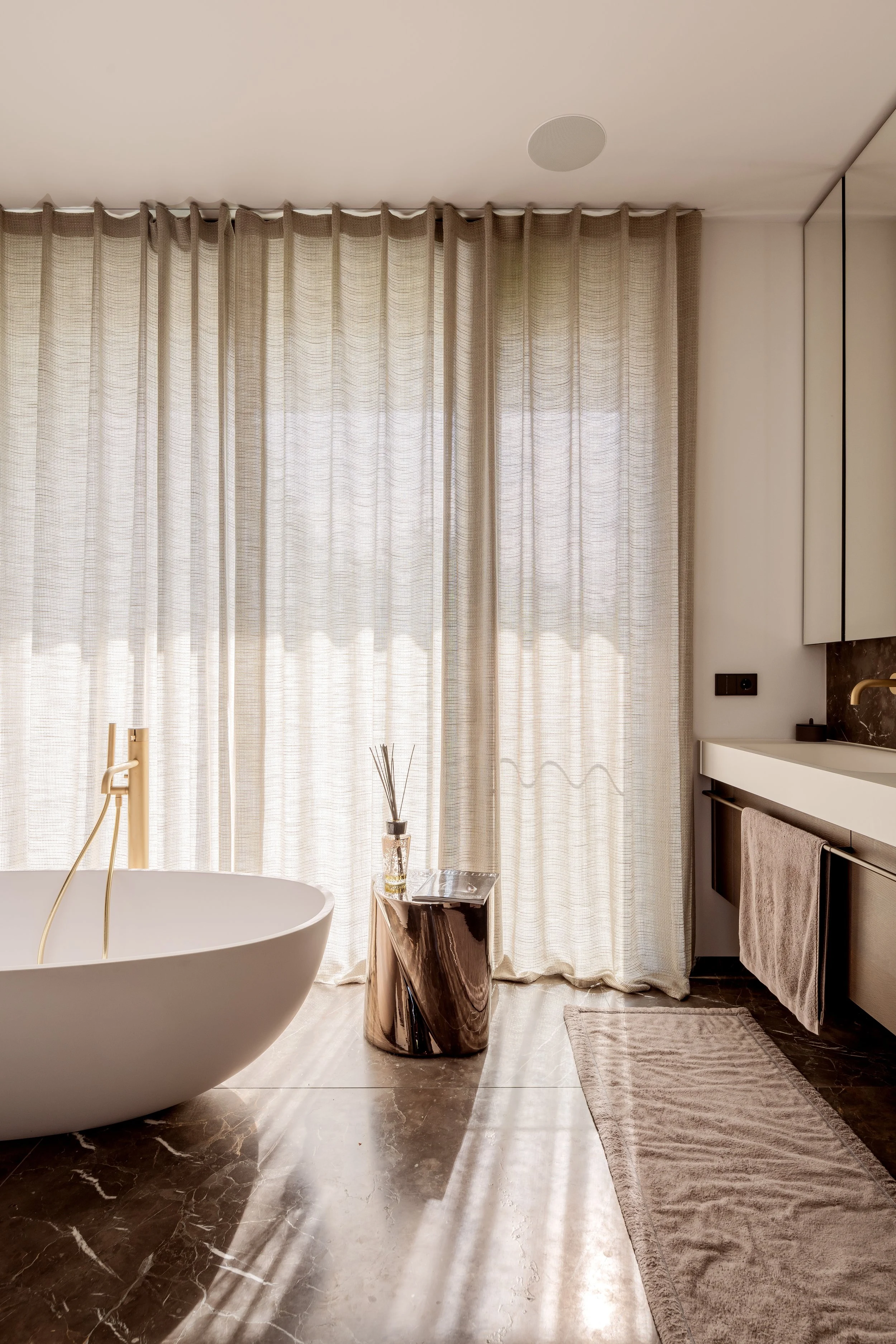 Ein Badezimmer mit einer freistehenden Badewanne, dunkelbraunem Fliesenboden, cremefarbenen Vorhängen, einem hellen Waschtisch mit Handtuch und einer kleinen silbernen Tisch mit Dekoration.