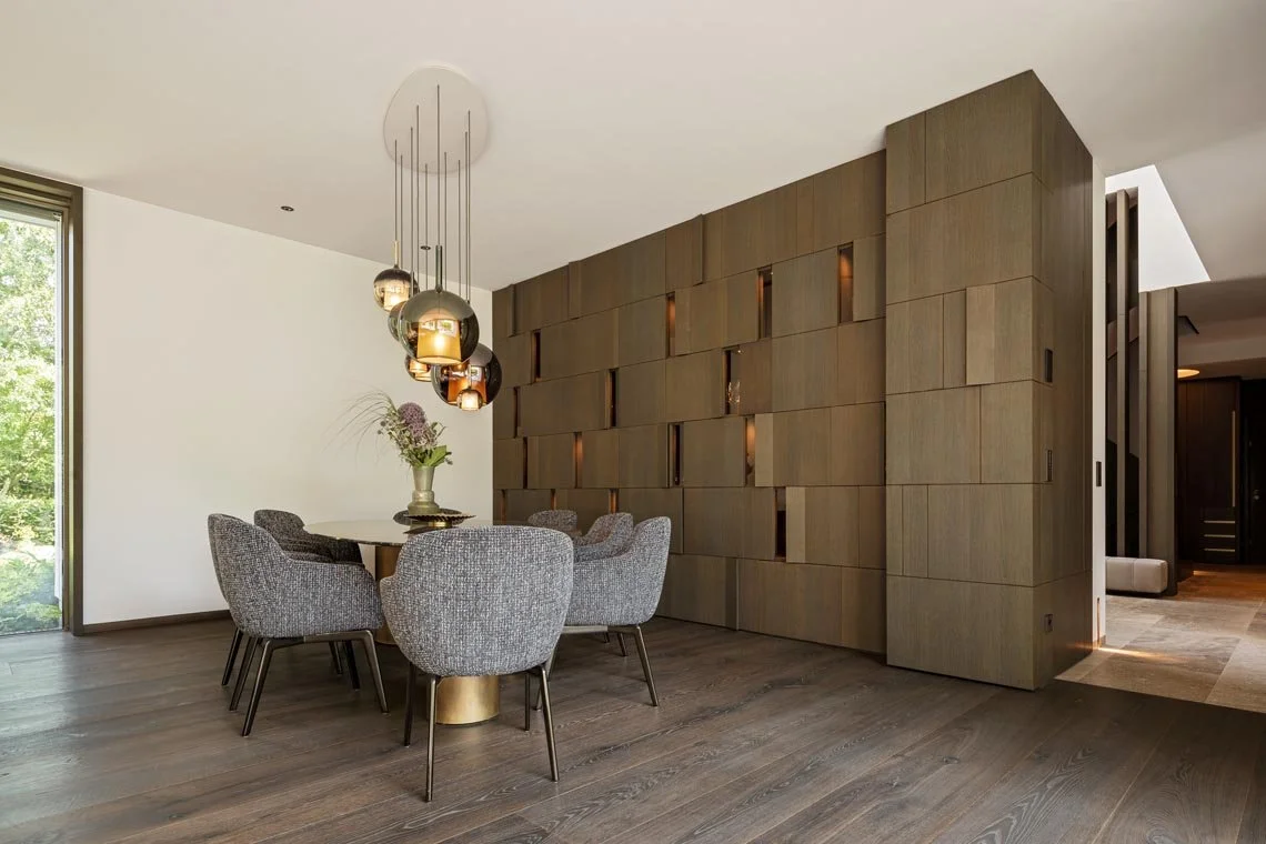 Moderner Essbereich mit einem runden Tisch, sechs grauen Polsterstühlen und einer stilvollen Hängeleuchte. Hintergrundwand aus Holzpaneelen mit integrierten Lichtern.