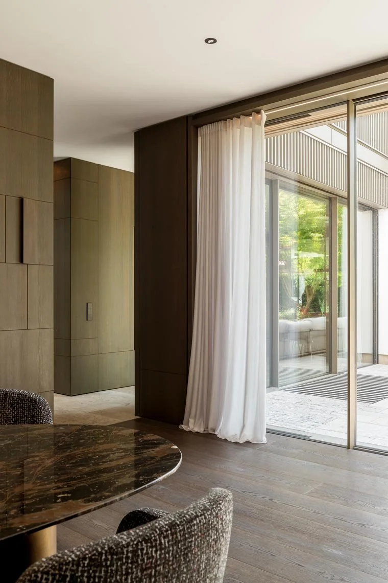 Innenaufnahme eines modernen Raumes mit Holzfußboden, einem runden dunklen Tisch und Stühlen. Große Glas-Schiebetüren führen auf einen Balkon mit Holzdecke und Grünblick. Weiße Vorhänge hängen vor den Türen.