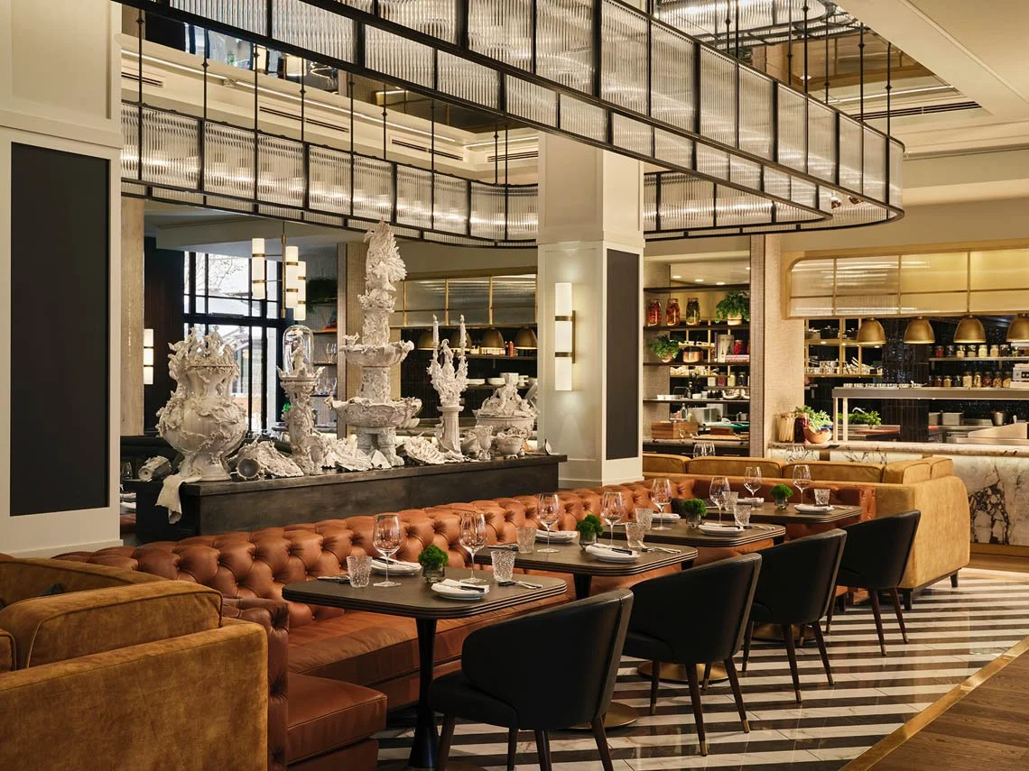 Elegantes Restaurant mit Tischen, Stühlen und dekorativen weißen Skulpturen auf einem schwarzen Podest, moderne Beleuchtung und eine offene Küchen- oder Bar-Ansicht im Hintergrund.