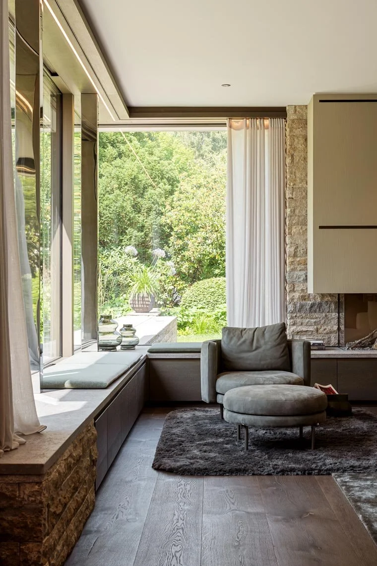 Ein modernes Wohnzimmer mit einer großen Fensterfront, weißen Vorhängen, einem grauen Sessel und einem schwarzen Teppich. Draußen ist ein Garten mit grünen Büschen und Bäumen sichtbar.