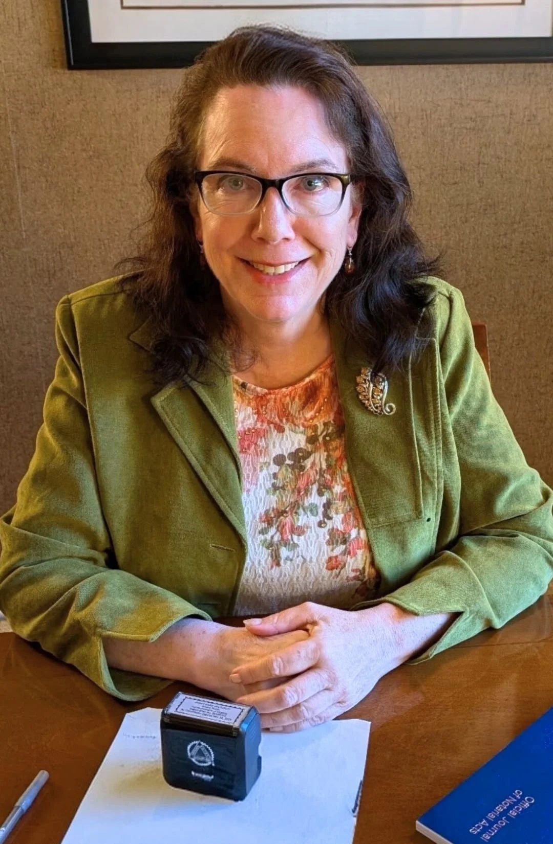 A woman with glasses and brown hair smiling, sitting at a table with a pen, paper, and a blue book. She is wearing a green jacket and a floral blouse, with a decorative brooch on her jacket.