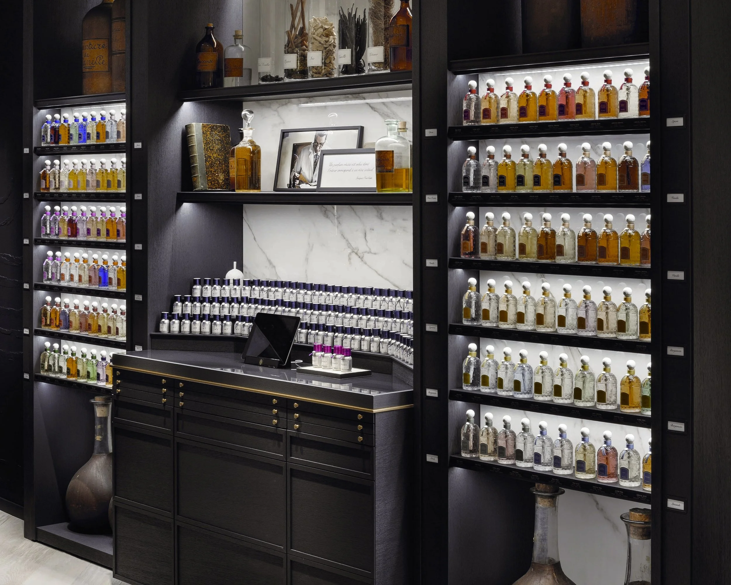 Guerlain boutique _ Étagères remplies de flacons de parfums colorés, devant un comptoir noir avec un miroir et quelques décorations