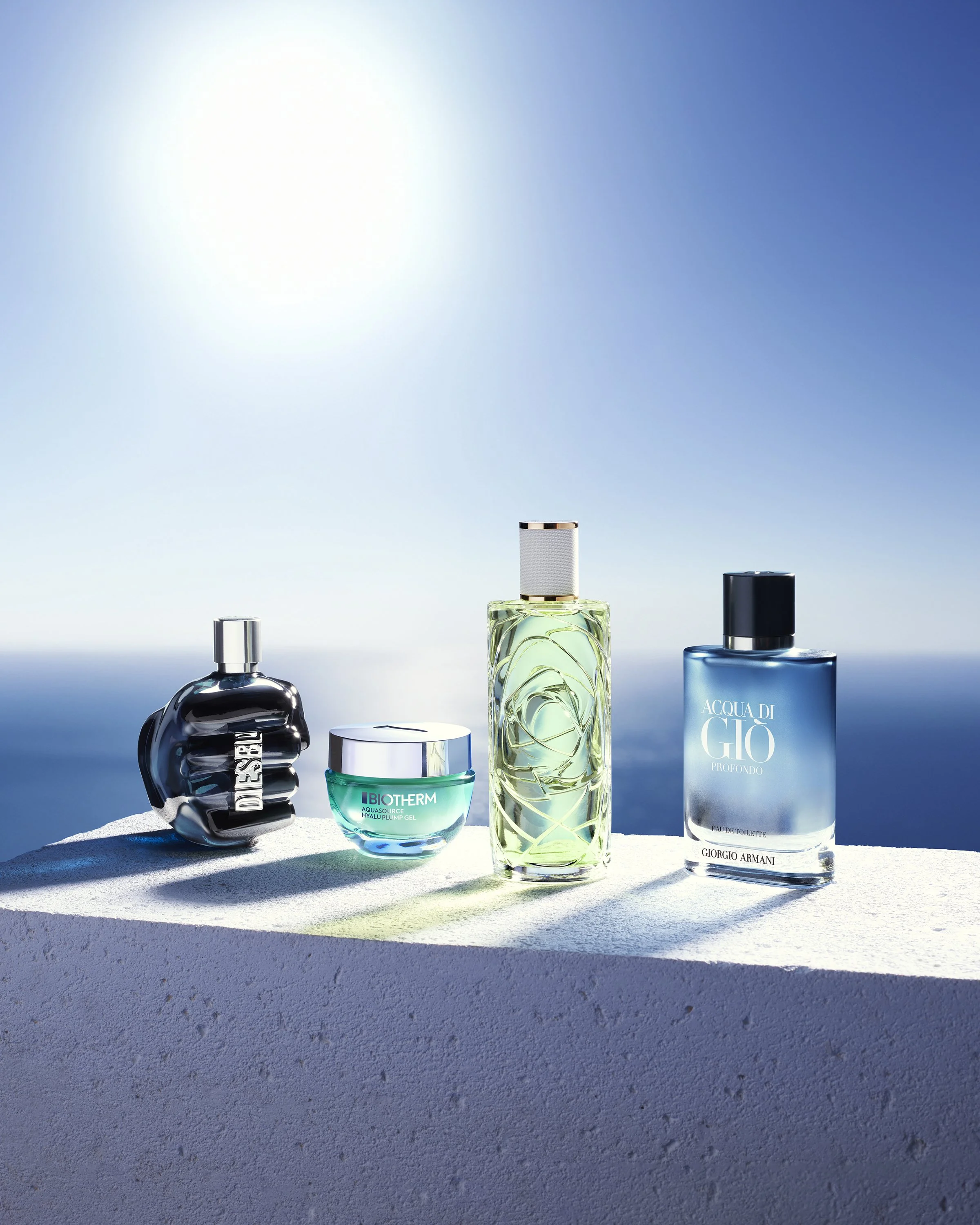 L'Oréal Luxe Summer Campaign _ Quatre flacons de parfum sur une surface blanche avec un fond de ciel bleu et mer. De gauche à droite : Diesel, Biotherm, un parfum non identifié, Giorgio Armani.