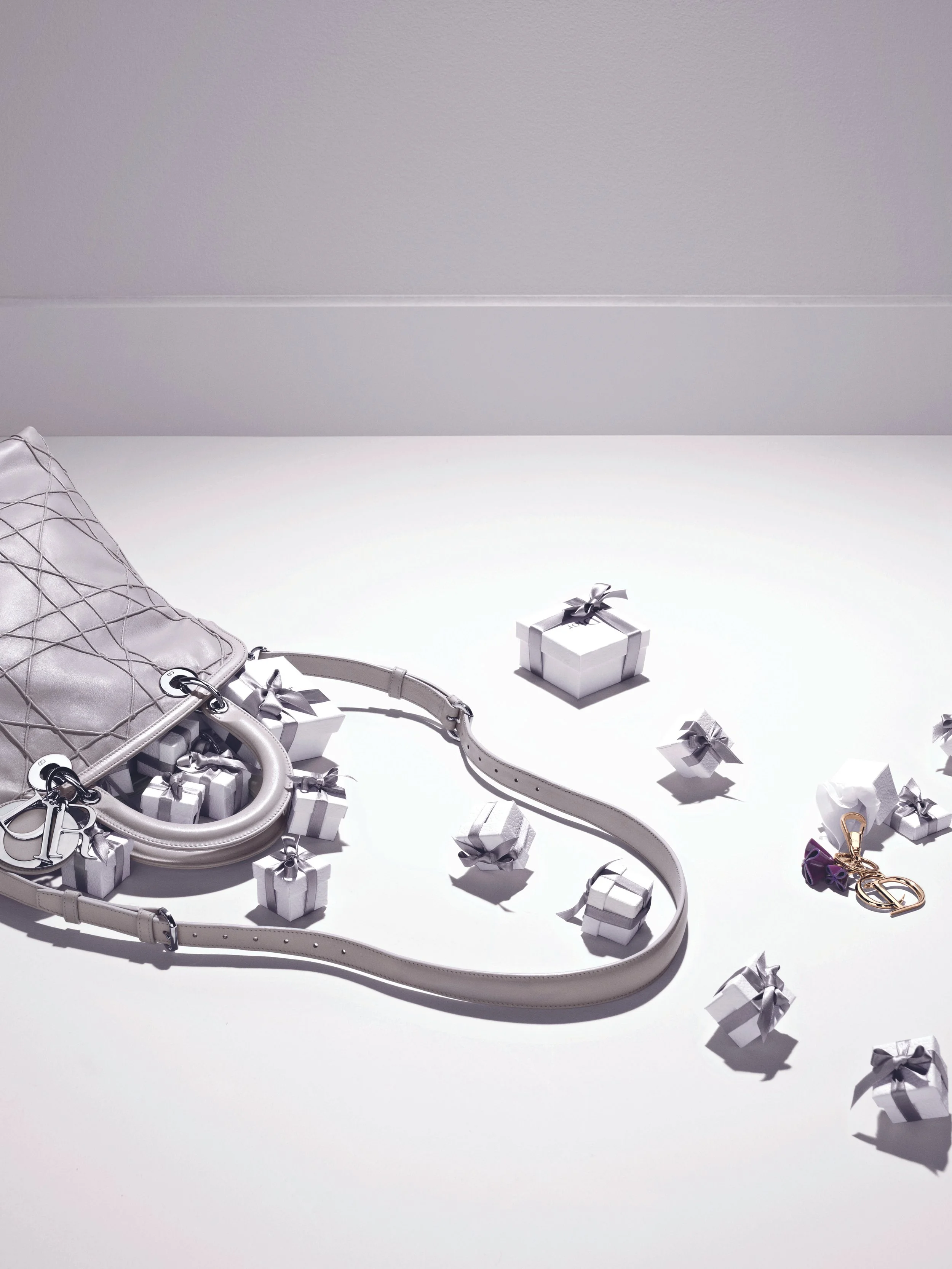 DIOR _ Sac à main blanc, petits cadeaux blancs avec rubans, clé en or, et porte-clés avec un cœur, disposés sur une table blanche.