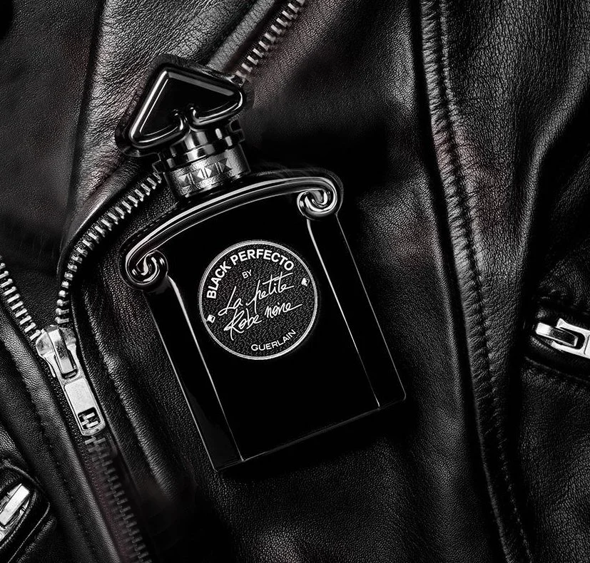 GUERLAIN Black Perfecto by La Petite Robe Noire _ Un flacon de parfum noir avec une étiquette indiquant "Black Perfecto Guerlain" placé sur une veste en cuir noire avec ferme zippée.