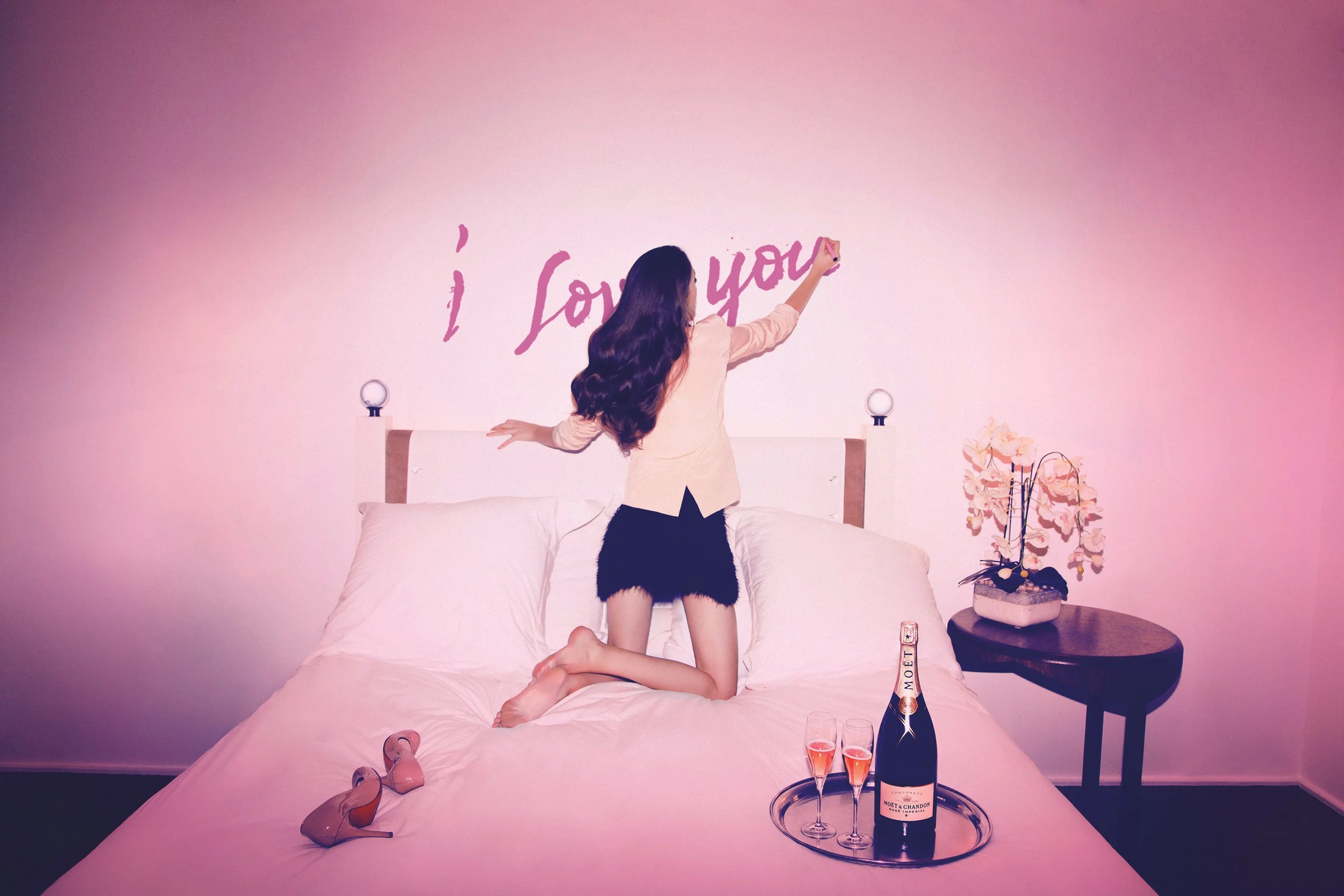 Moët&Chandon Collab André _ Femme en tailleur écrivant 'I Love You' sur un mur rose dans une chambre, avec une bouteille de champagne, deux flûtes, une paire de chaussures à talons roses, et une plante fleurie sur une table