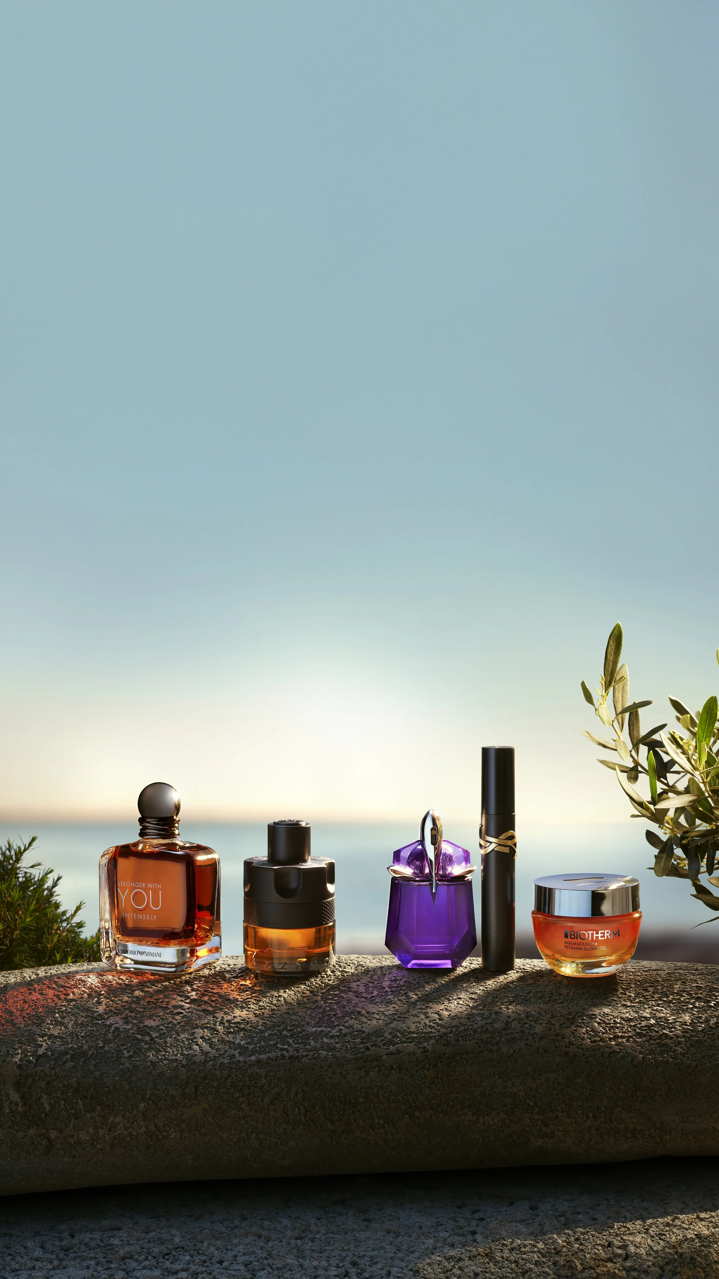 L'Oréal Luxe Summer campaign _ Collection de cinq flacons de parfum posés sur une roche avec un fond de ciel bleu et des végétaux à droite. Les flacons varient en forme et en couleur, comprenant des étuis noir, violet et orange.