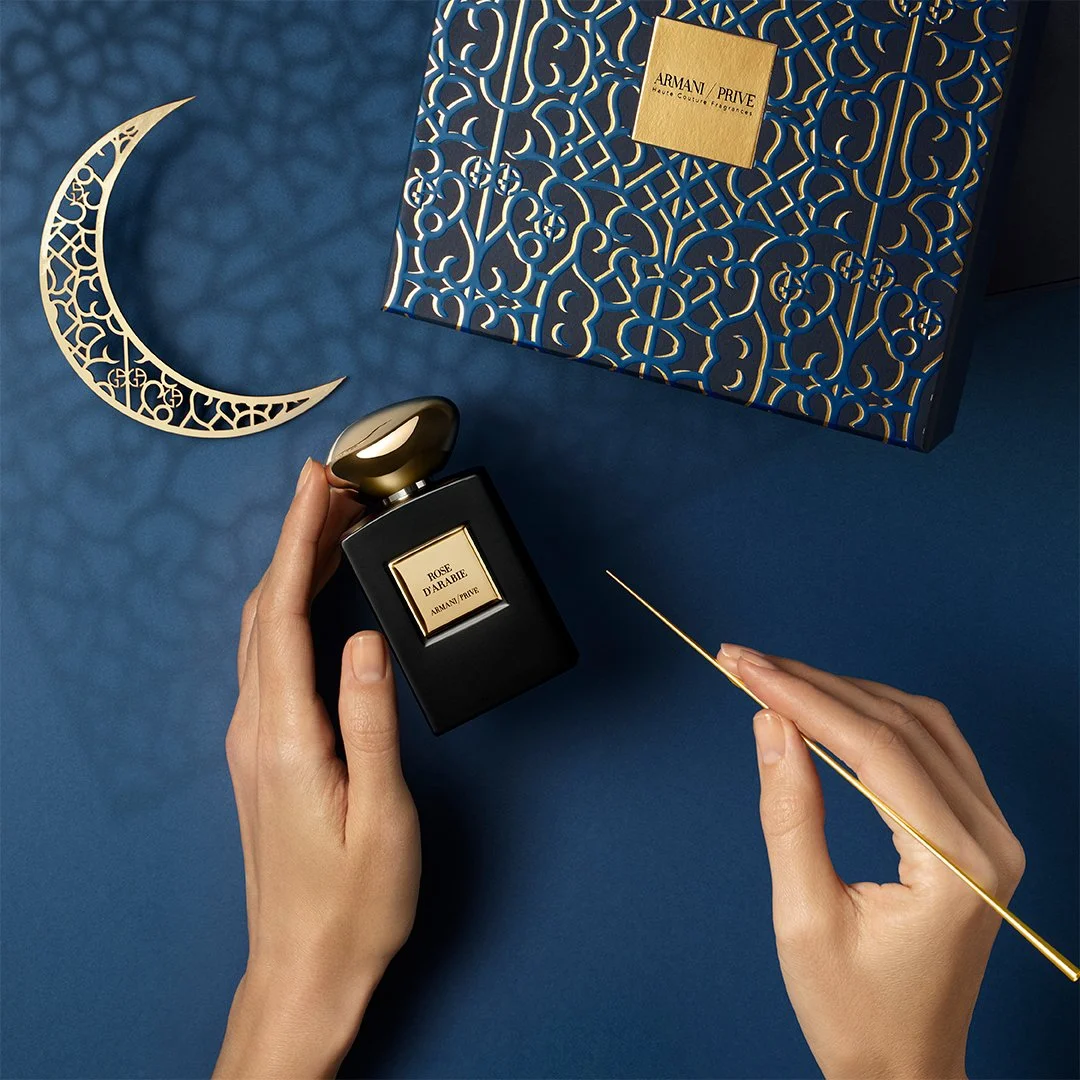 Un flacon de parfum noir avec un bouchon doré, intitulé « Rose d'Arabie » de la collection Armani Privé, est tenu dans une main. Une aiguille en or est tenue dans l'autre main. Un coffret bleu et or orné de motifs géométriques est visible à l'arrière. Une décoration en forme de croissant de lune doré est également présente.