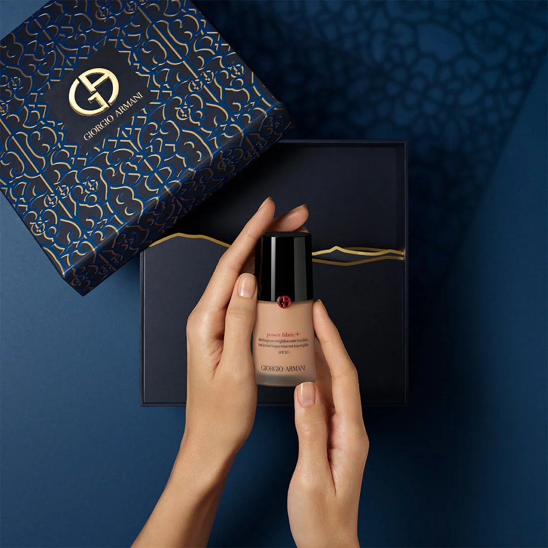 ARMANI Ramadan Gifting _ Une main tient un flacon de fond de teint Giorgio Armani dans une boîte noire, avec une boîte décorative en bleu et or à côté, sur un fond bleu.