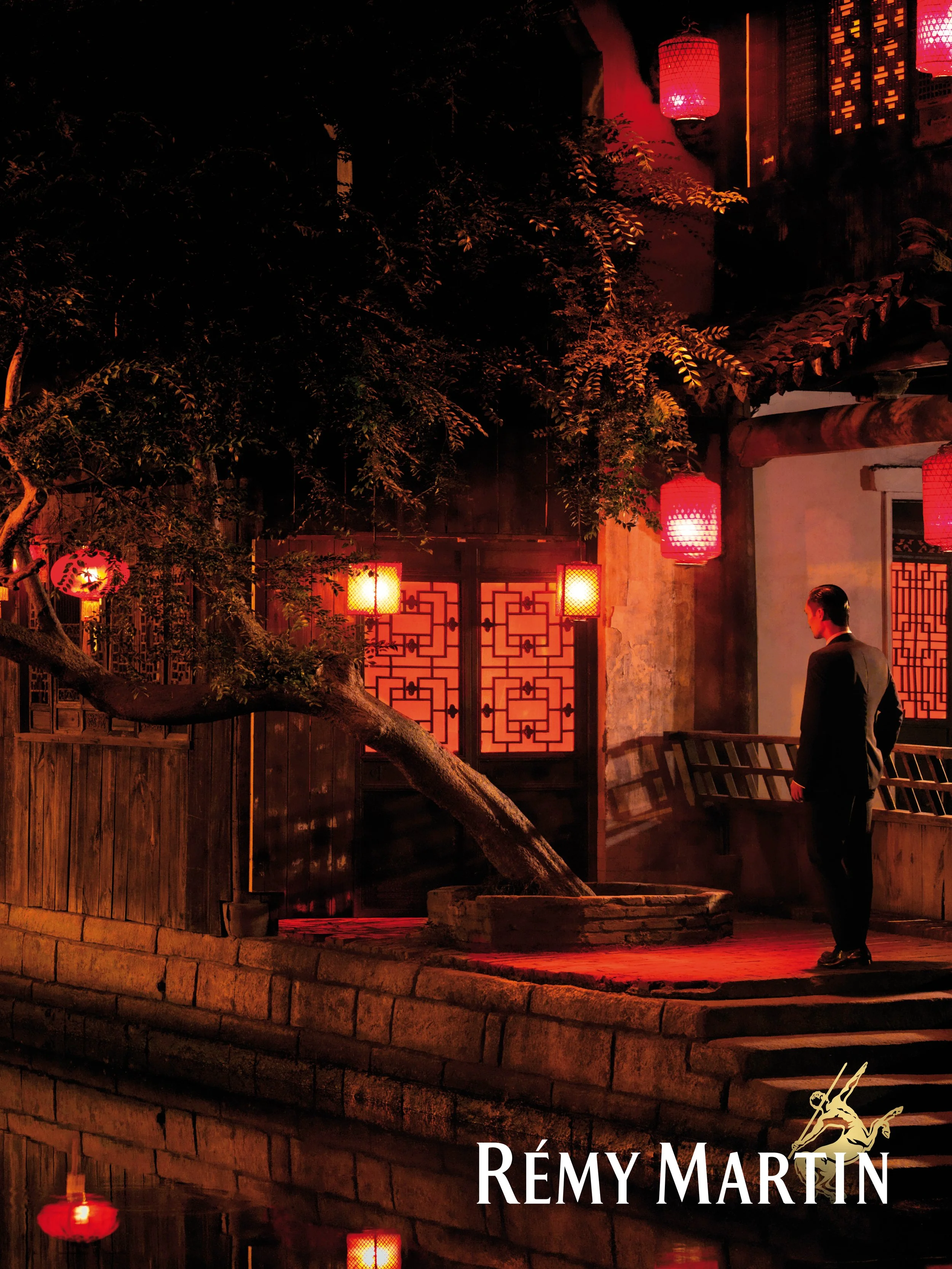 Rémy Martin Coupe Shanghaï Campaign_Une scène nocturne dans un cadre asiatique avec des lanternes rouges suspendues, un homme en costume noir se tenant sur un pont en pierre