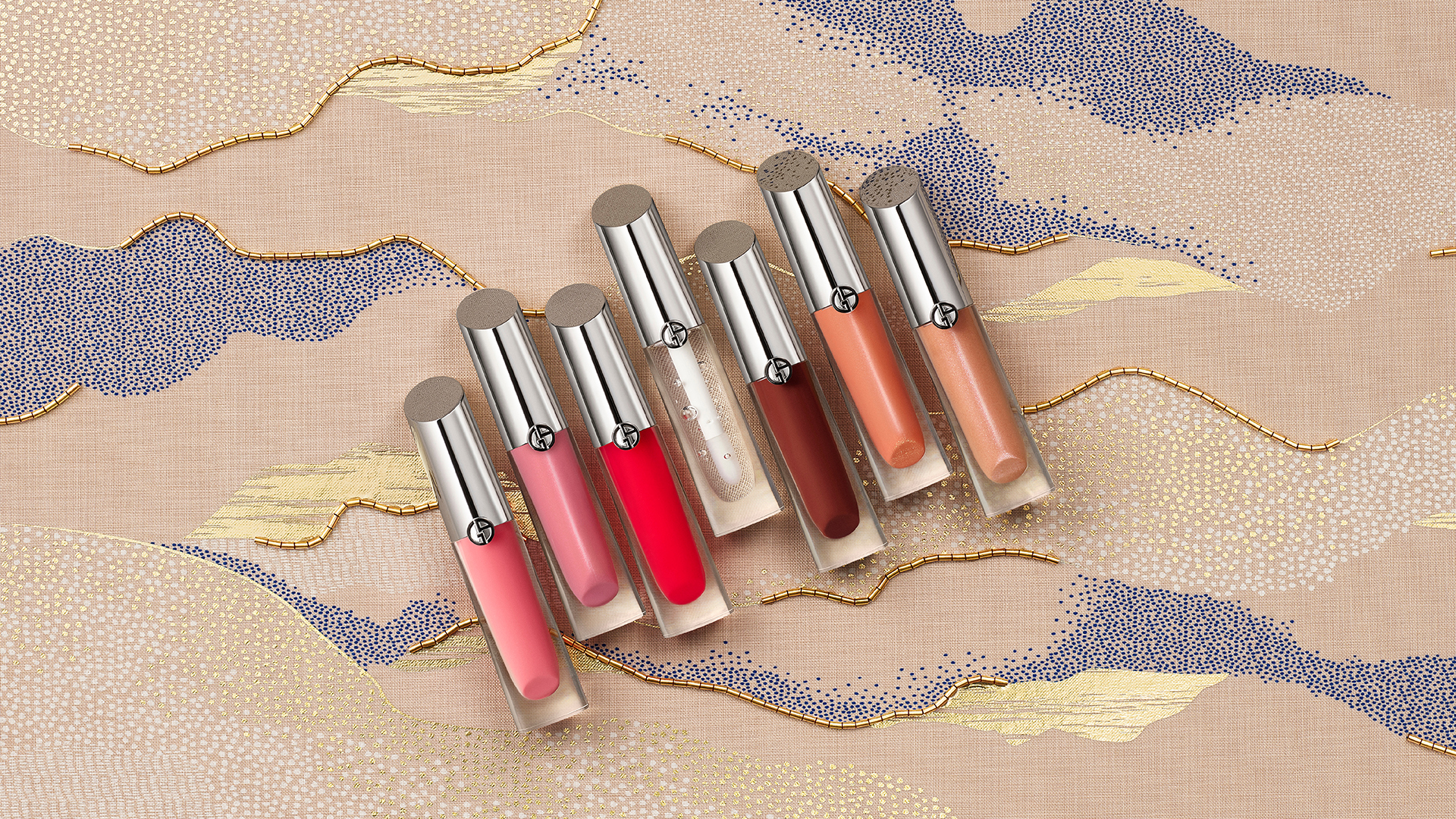 ARMANI Ramadan _ Sept tubes de gloss à lèvres Prisma Glass, alignés sur un tissu décoré avec des motifs perlés représentant de manière abstraite des dunes de sable, le désert