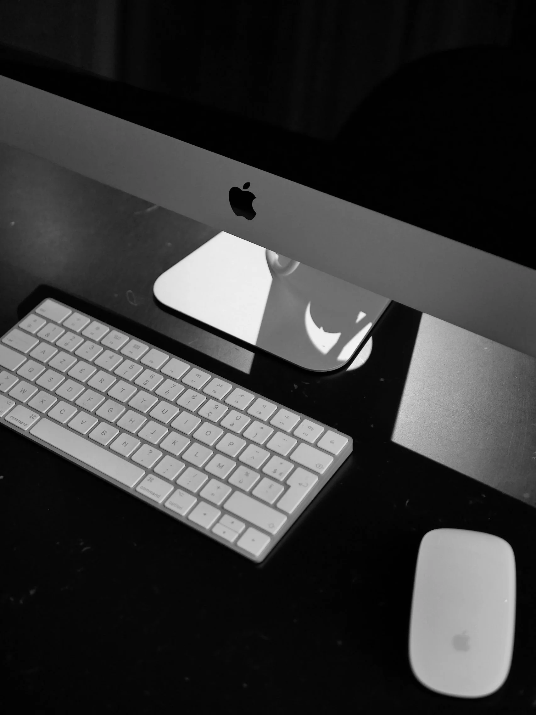 Écran d'ordinateur Apple, clavier et souris sur un bureau noir, photographie en noir et blanc.