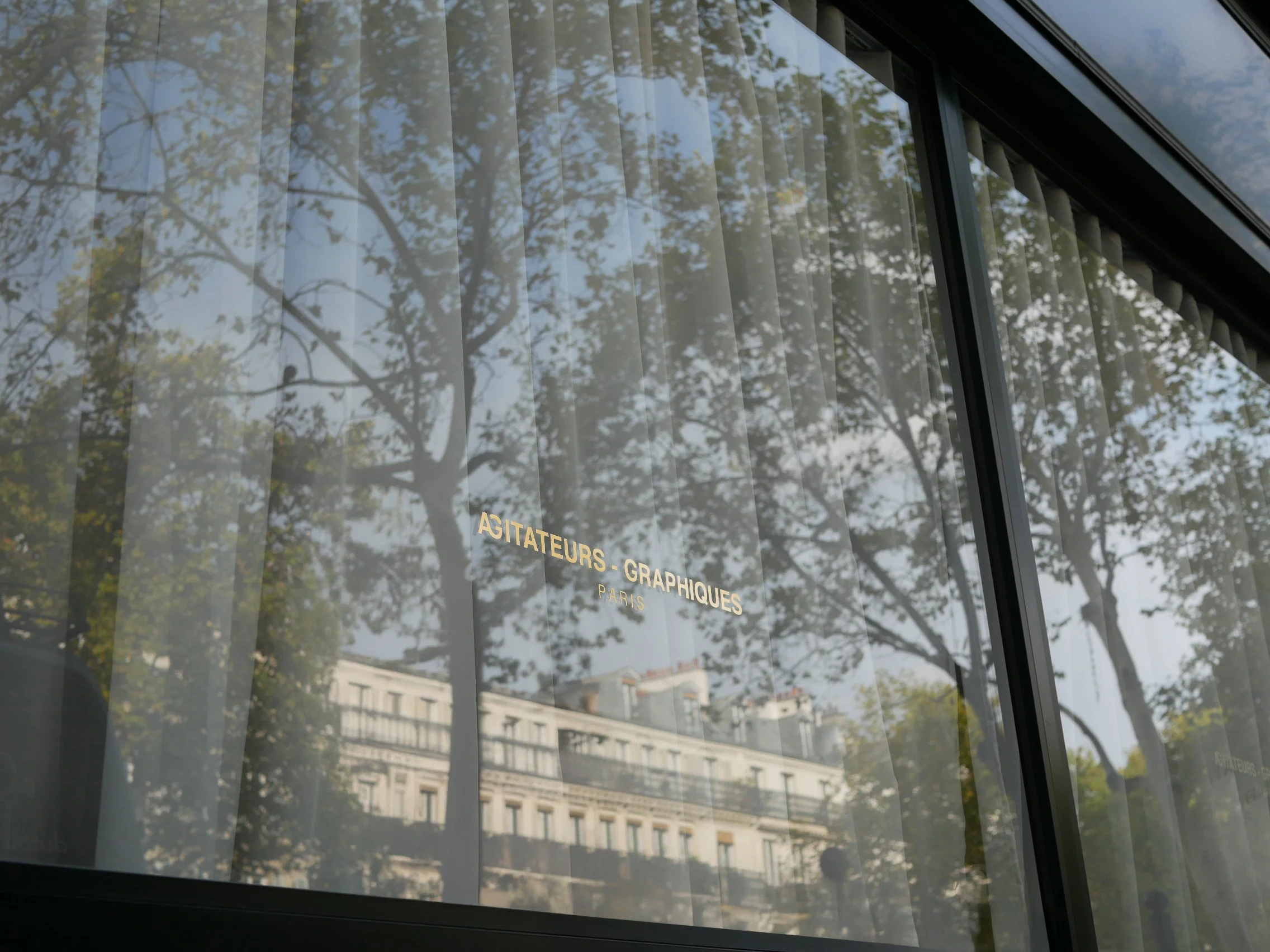 Vitrine avec l'inscription 'Agitateurs Graphiques Paris' reflétant des arbres et un bâtiment.
