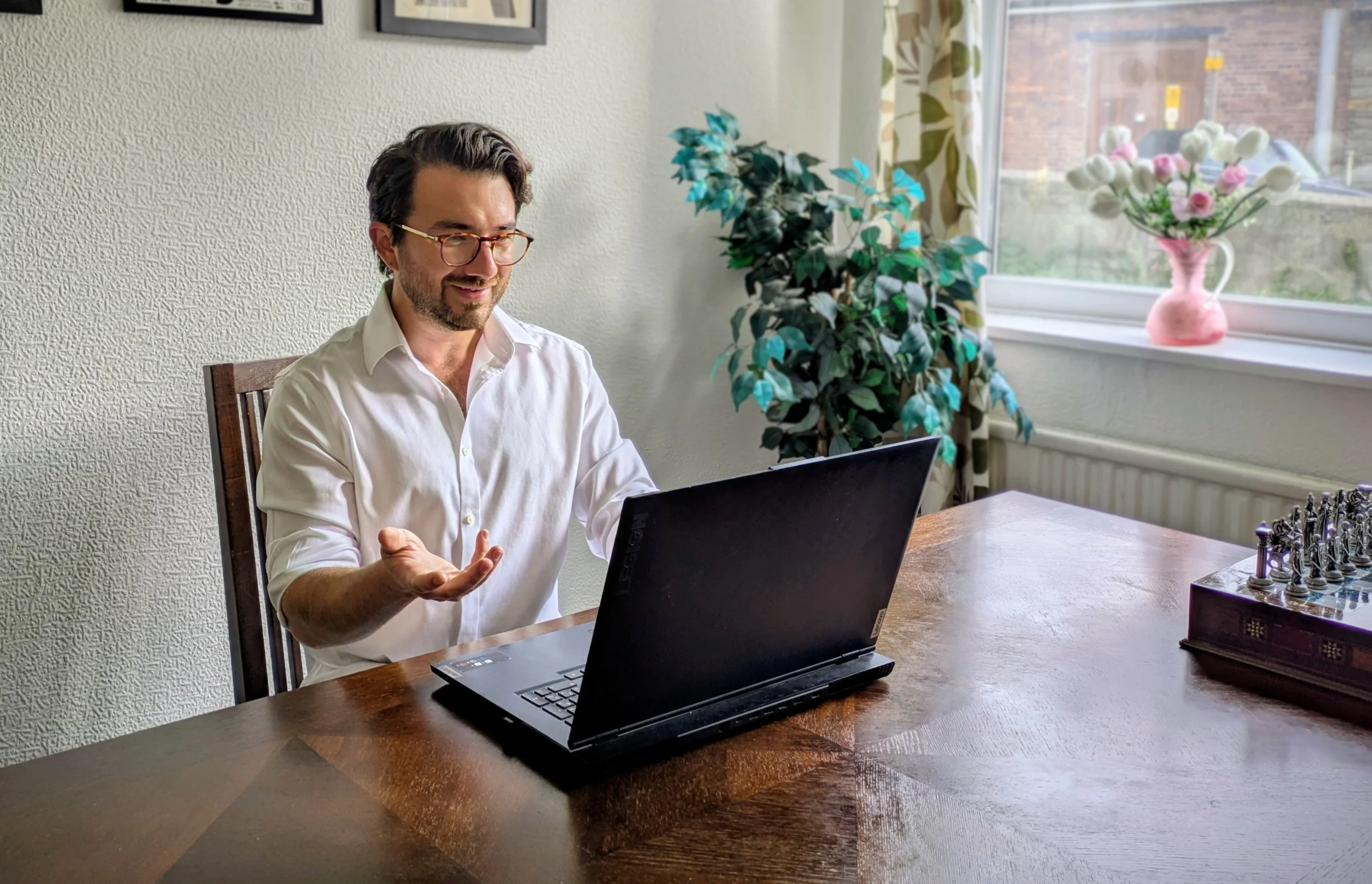 A man with glasses sitting at a wooden table using a black laptop, with a pink vase with flowers on the windowsill and a potted plant next to him.