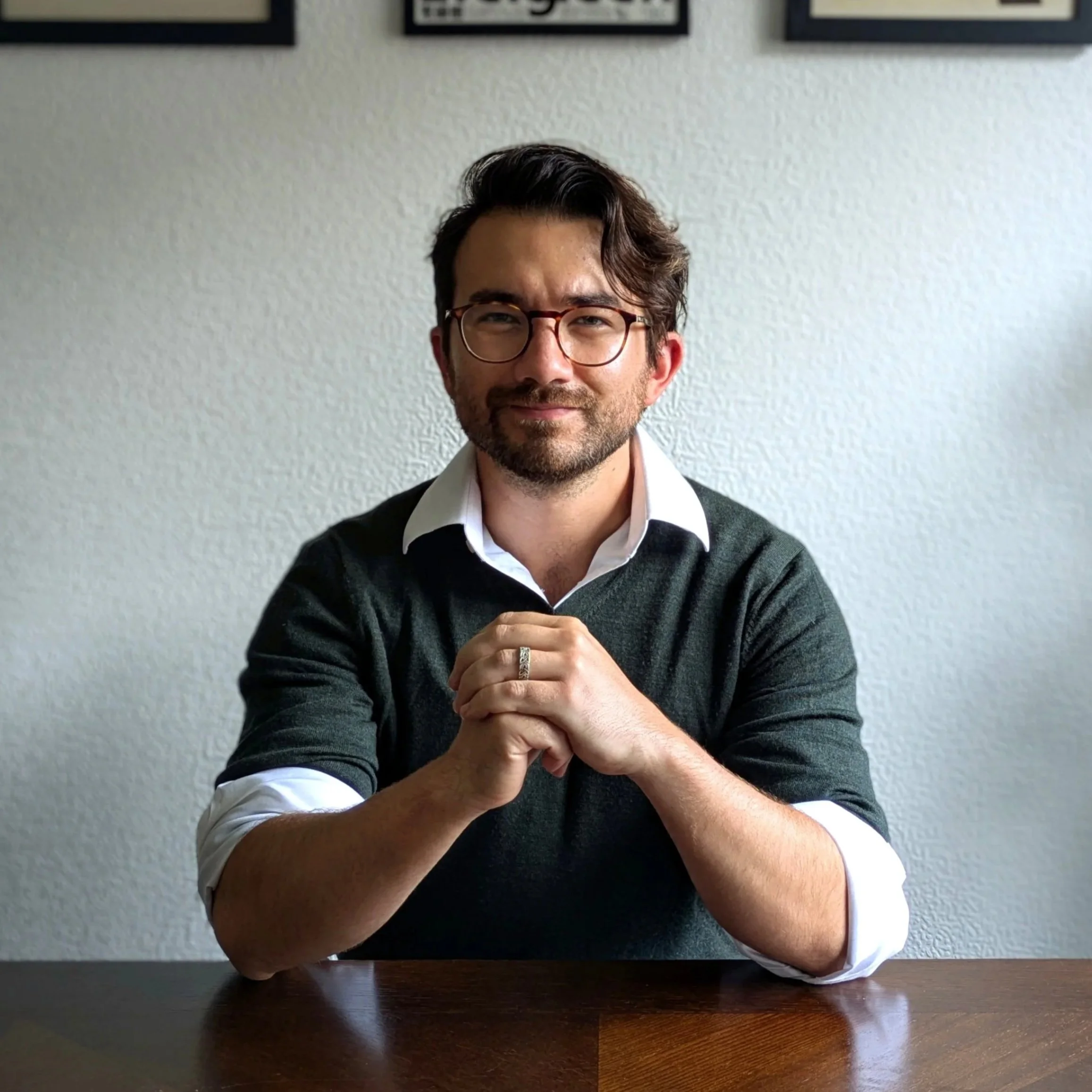 Photo of Liam: A man with glasses and a beard, wearing a dark sweater over a white shirt, sitting at a wooden table with his hands clasped, looking at the camera with a slight smile, in front of a plain light-colored wall.
