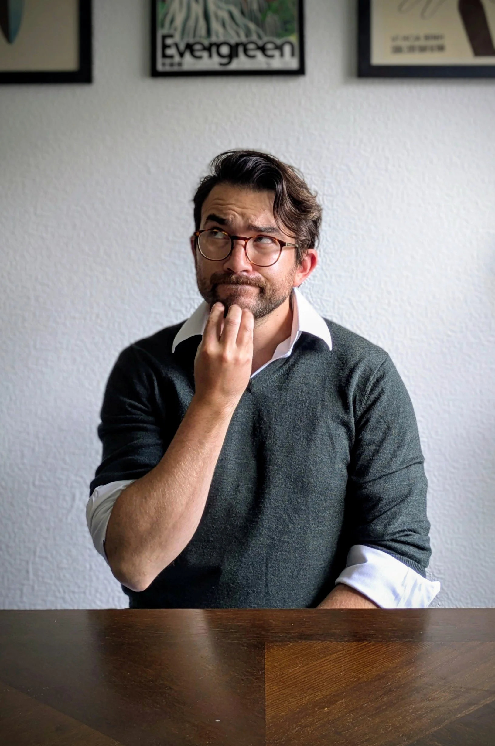 Photo of Liam: A man with glasses and dark hair, wearing a white collared shirt under a dark sweater, sits at a wooden table with a curious or contemplative expression, hand on chin, in front of a plain wall with framed artwork.
