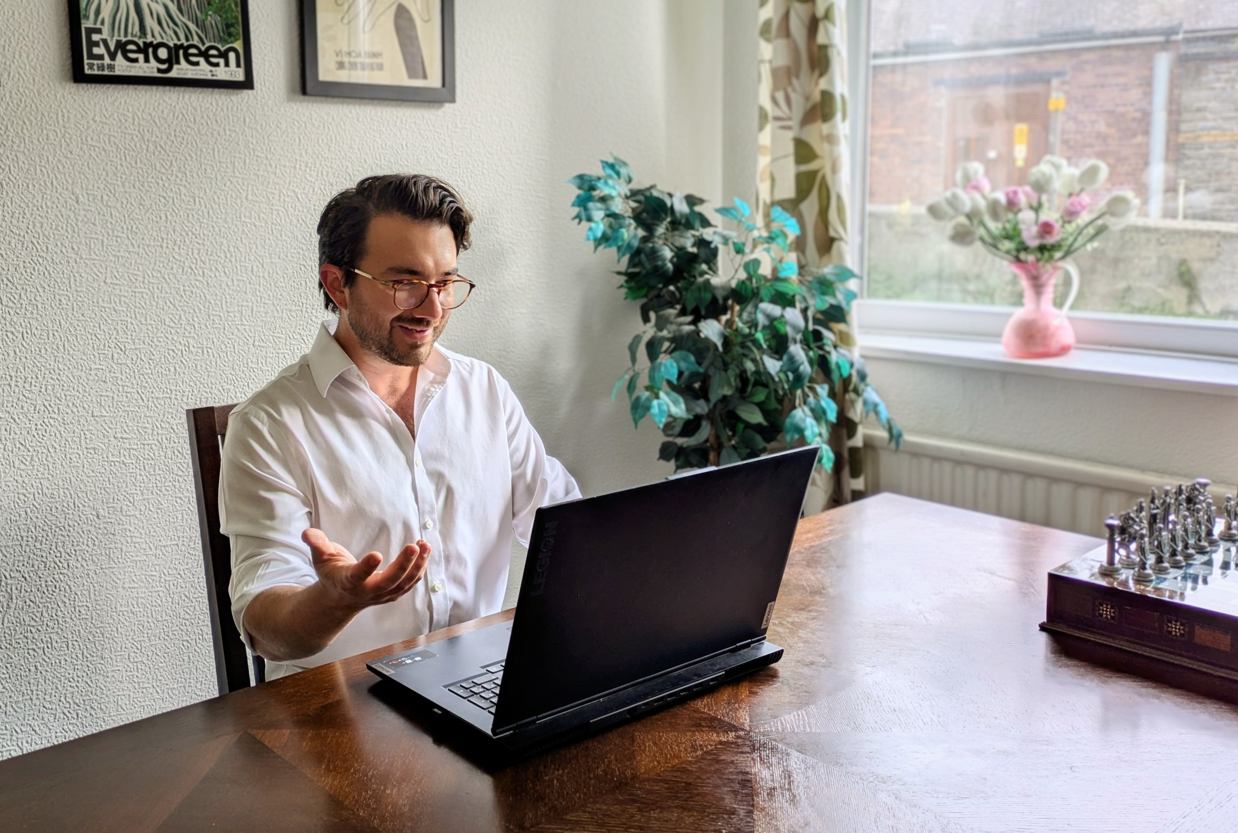 A man with dark hair and glasses sitting at a wooden table, using a black laptop, gesturing with his right hand and smiling