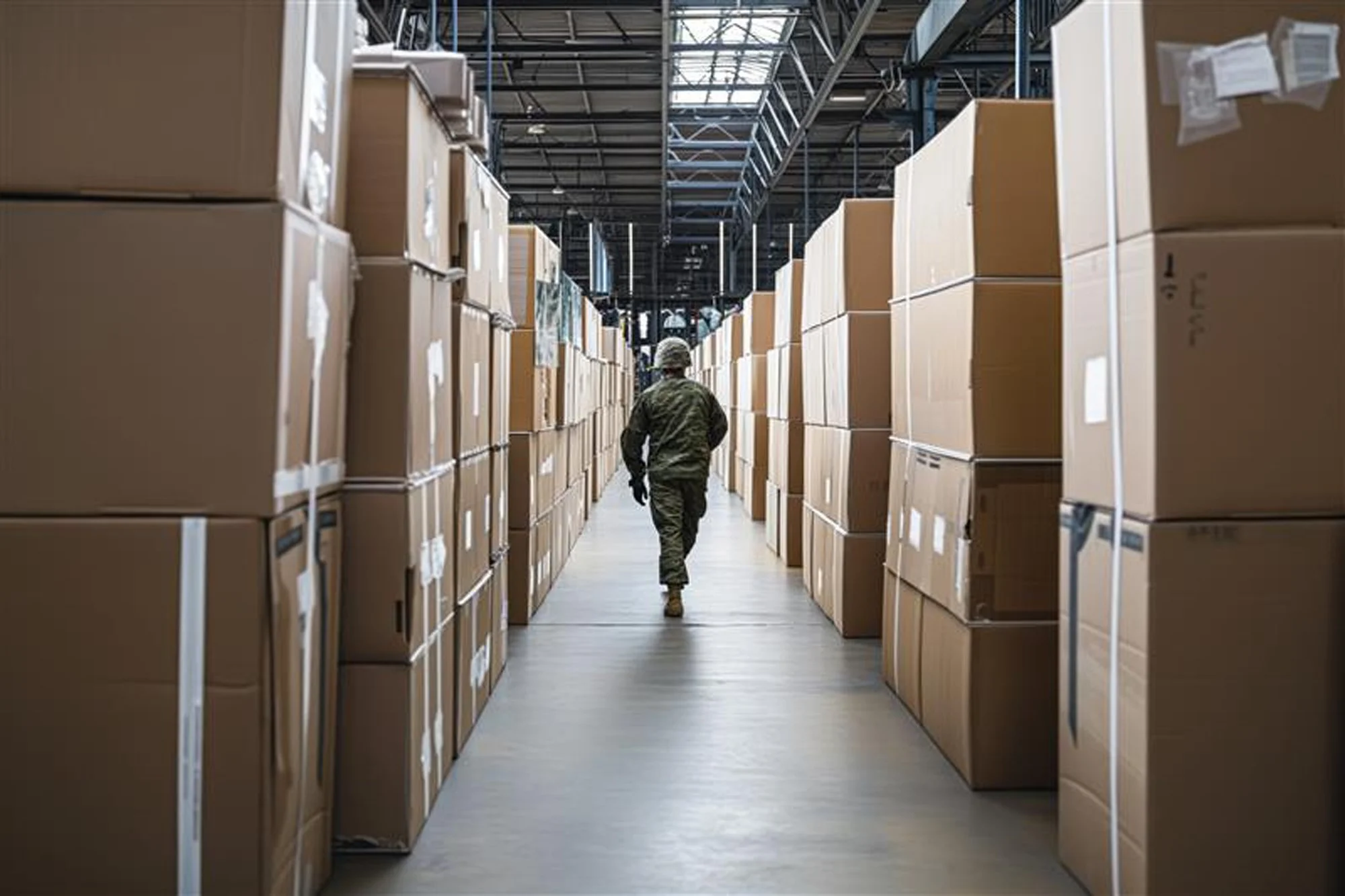 A person in military camouflage walk down an aisle in a warehouse with stacked cardboard boxes on both sides.