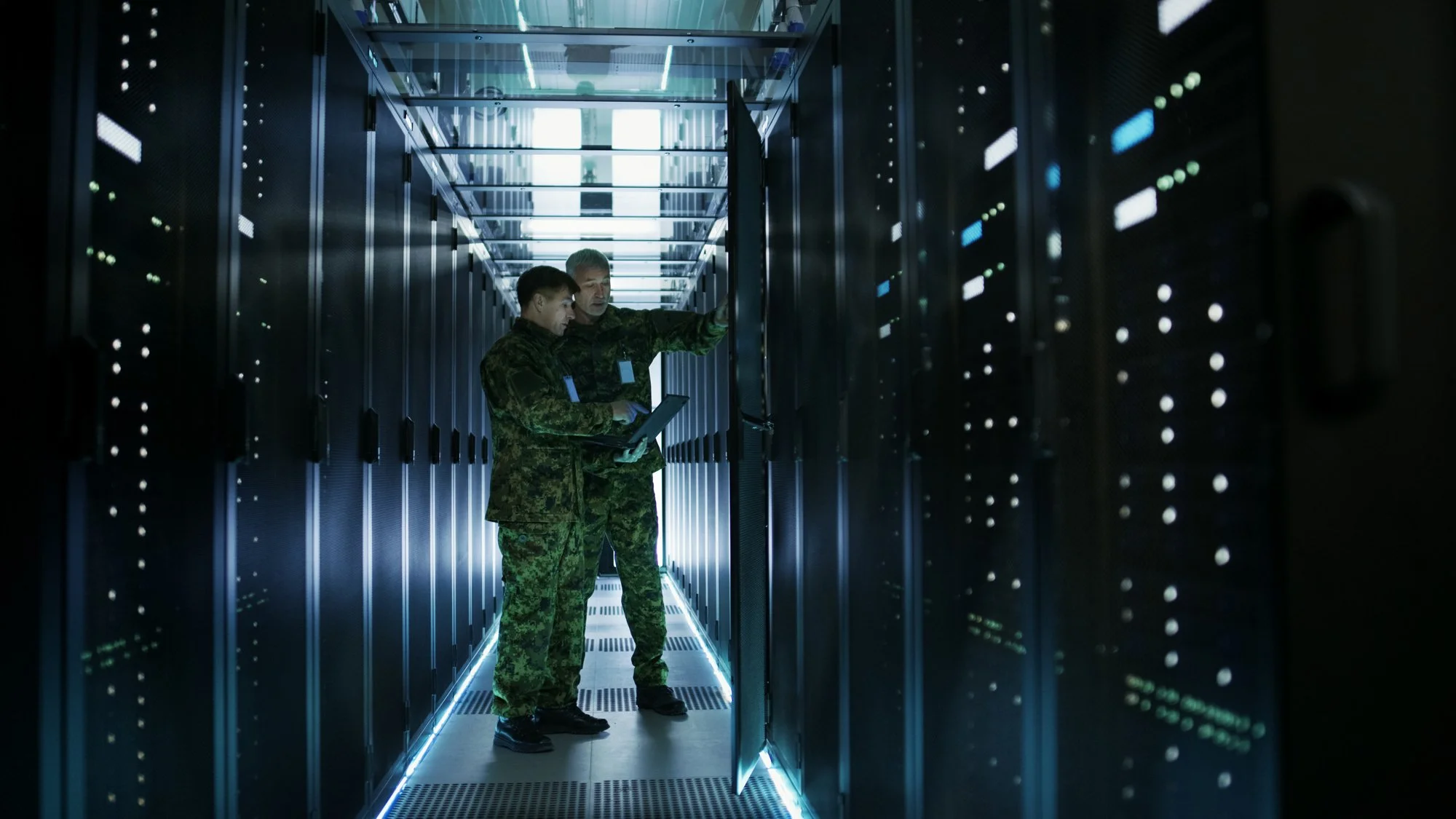 Two military personnel in camouflage uniforms inspecting servers in a data center.