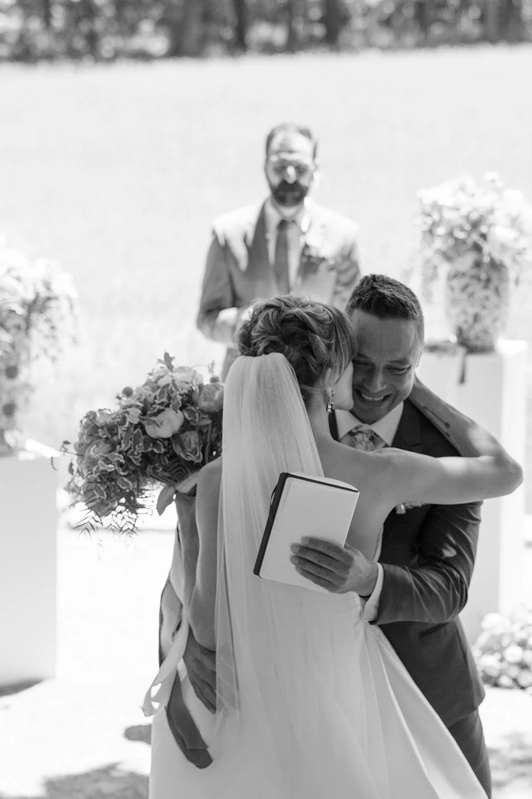 A bride and groom embrace during their outdoor wedding ceremony, with the officiant standing behind them.