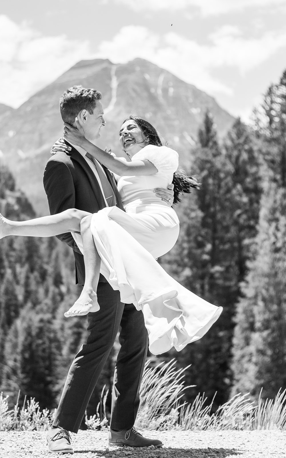 A man in a suit holding a woman in a wedding dress, smiling and looking into each other's eyes outdoors with mountains and trees in the background.