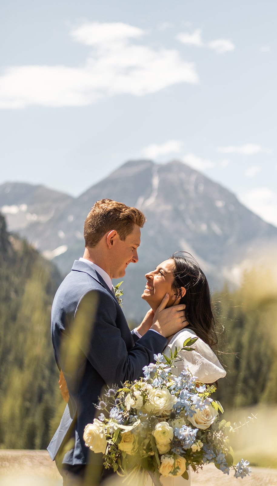 A newlywed couple sharing a joyful moment outdoors with mountains and blue sky in the background, surrounded by greenery.