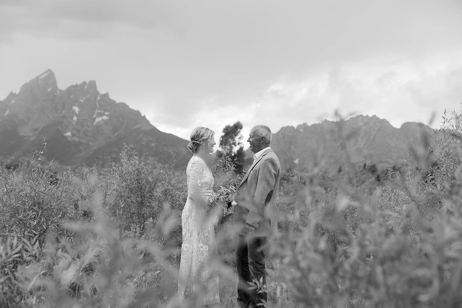 A black and white photo of a bride and groom standing face to face outdoors, surrounded by bushes, with mountains in the background.