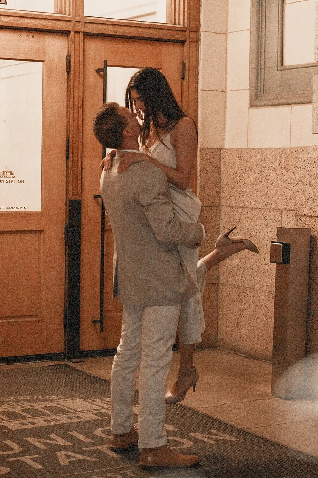 A man lifting a woman in a beige suit inside a building with wooden doors and walls.