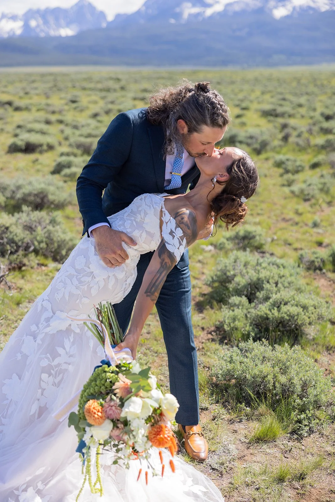 A newlywed couple kissing outdoors in a field, the man in a dark suit and the woman in a white wedding dress holding a bouquet of flowers, with mountains in the background.