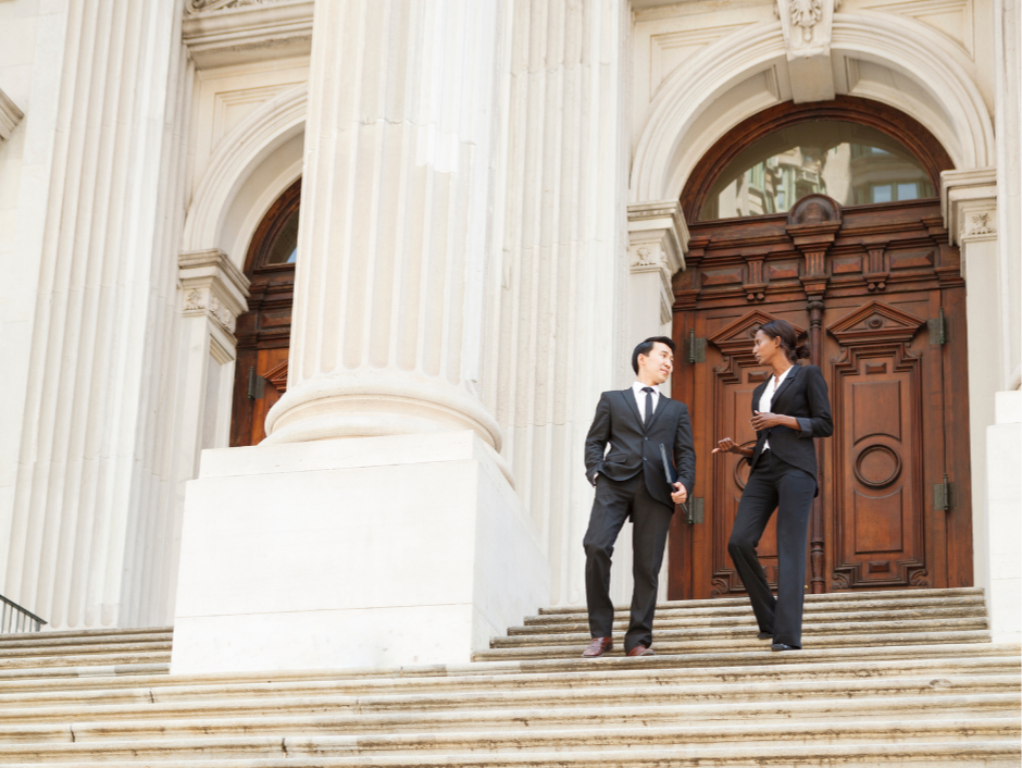 Two business professionals, a man and a woman, standing on steps outside a historic building with large white columns and wooden doors, engaged in conversation.